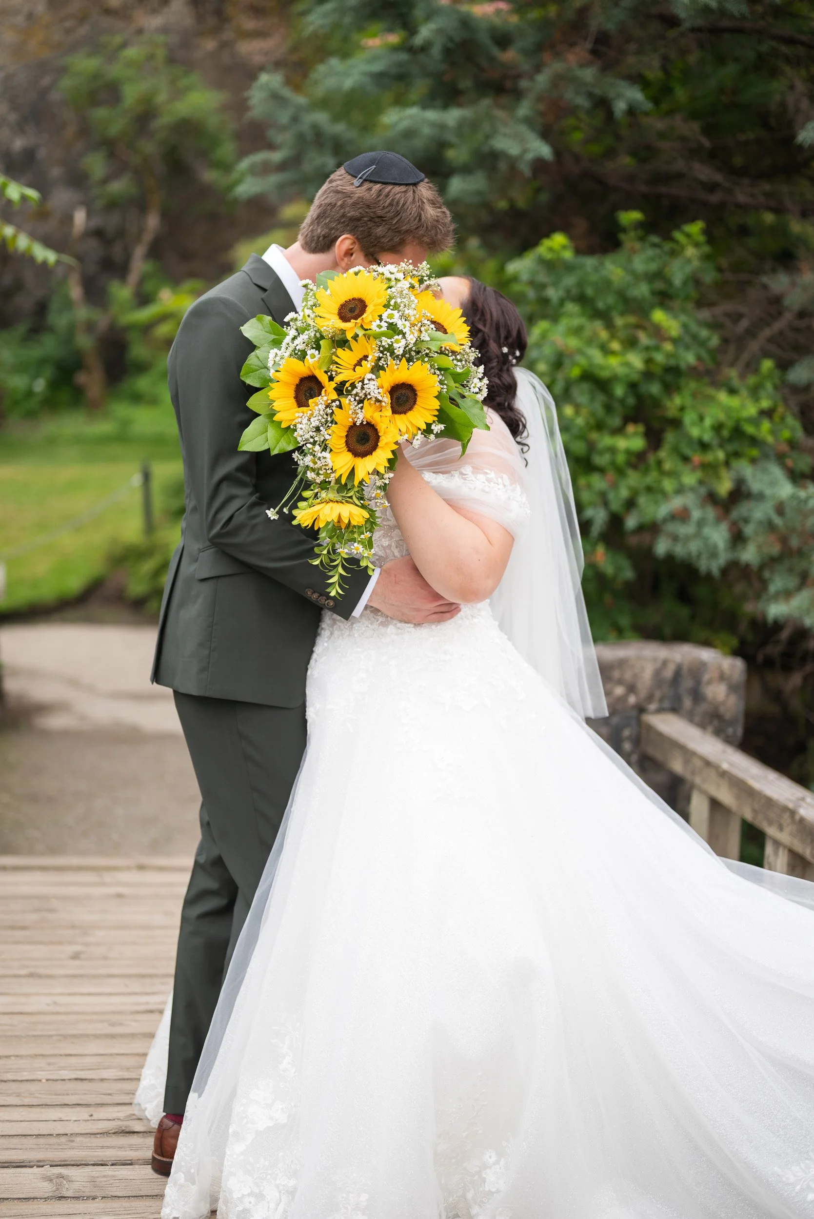 A bride and groom embrace and kiss outdoors on a wooden bridge at Queen Elizabeth Park, with the groom holding a large bouquet of sunflowers and white flowers.