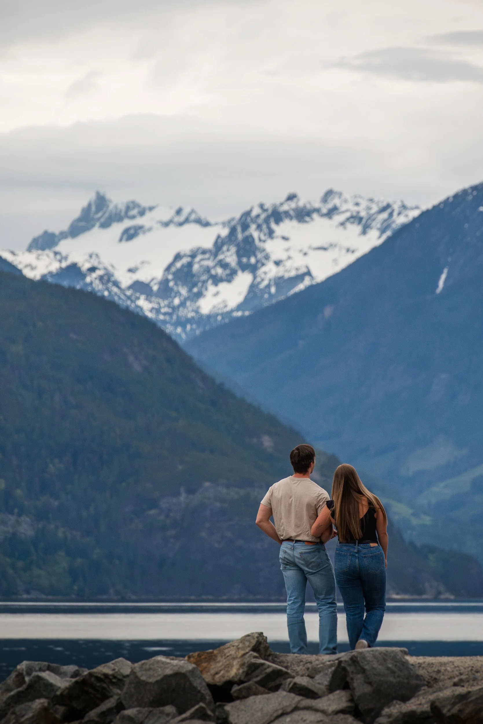 A couple standing on rocks near a body of water, overlooking snow-capped mountains with a cloudy sky.