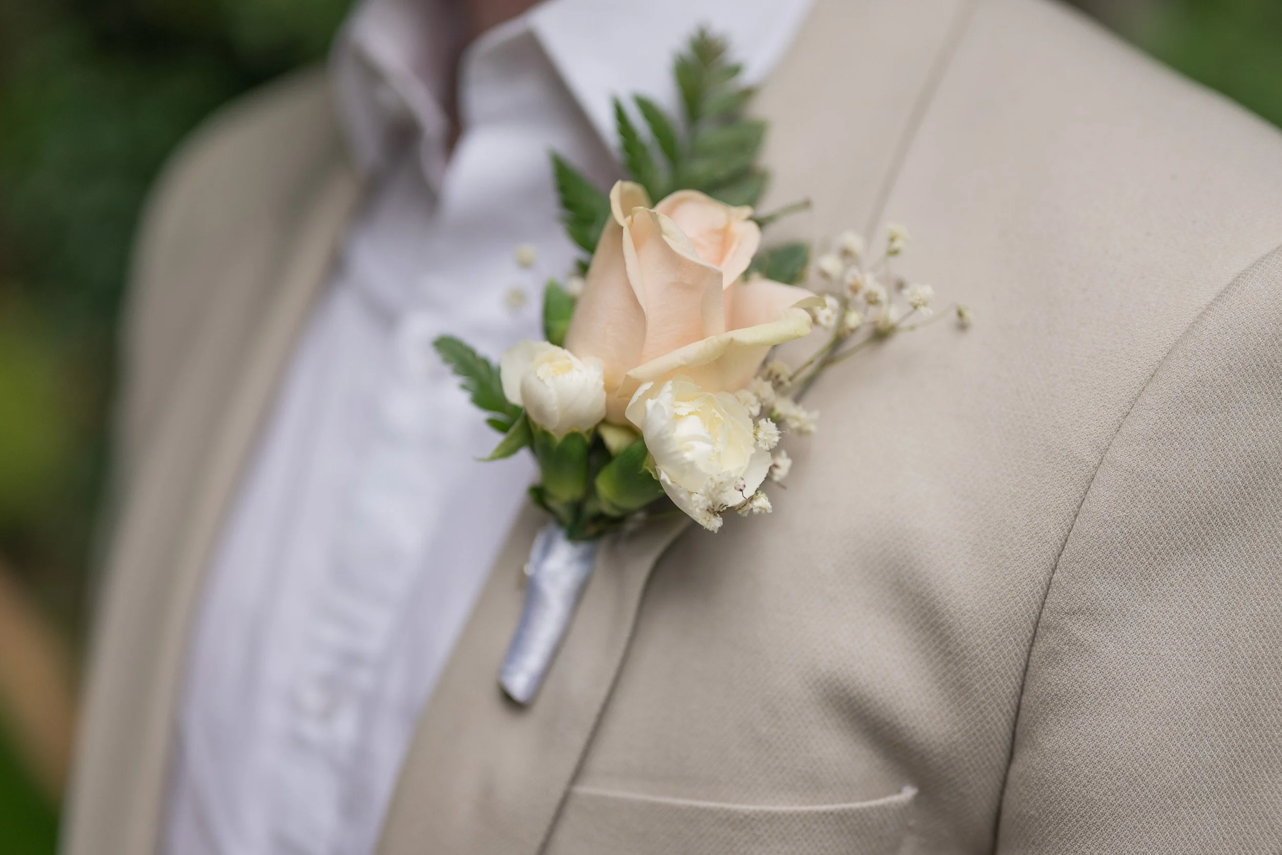 Close-up of a boutonniere with a light peach rose, white filler flowers, and green leaves, pinned to a beige suit lapel for a wedding.