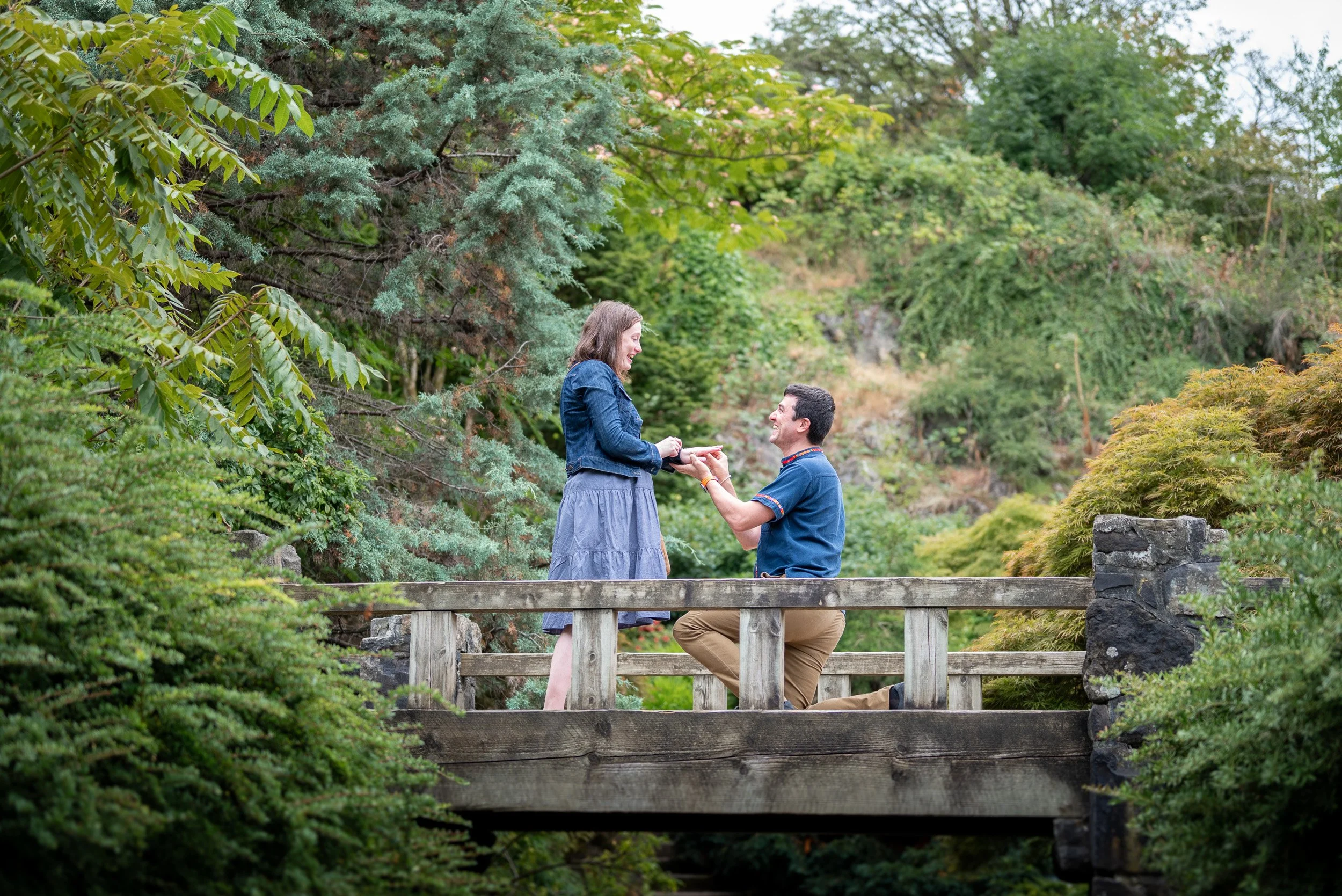 A man kneeling on one knee proposing to a woman on a bridge in a lush green garden at Queen Elizabeth Park.