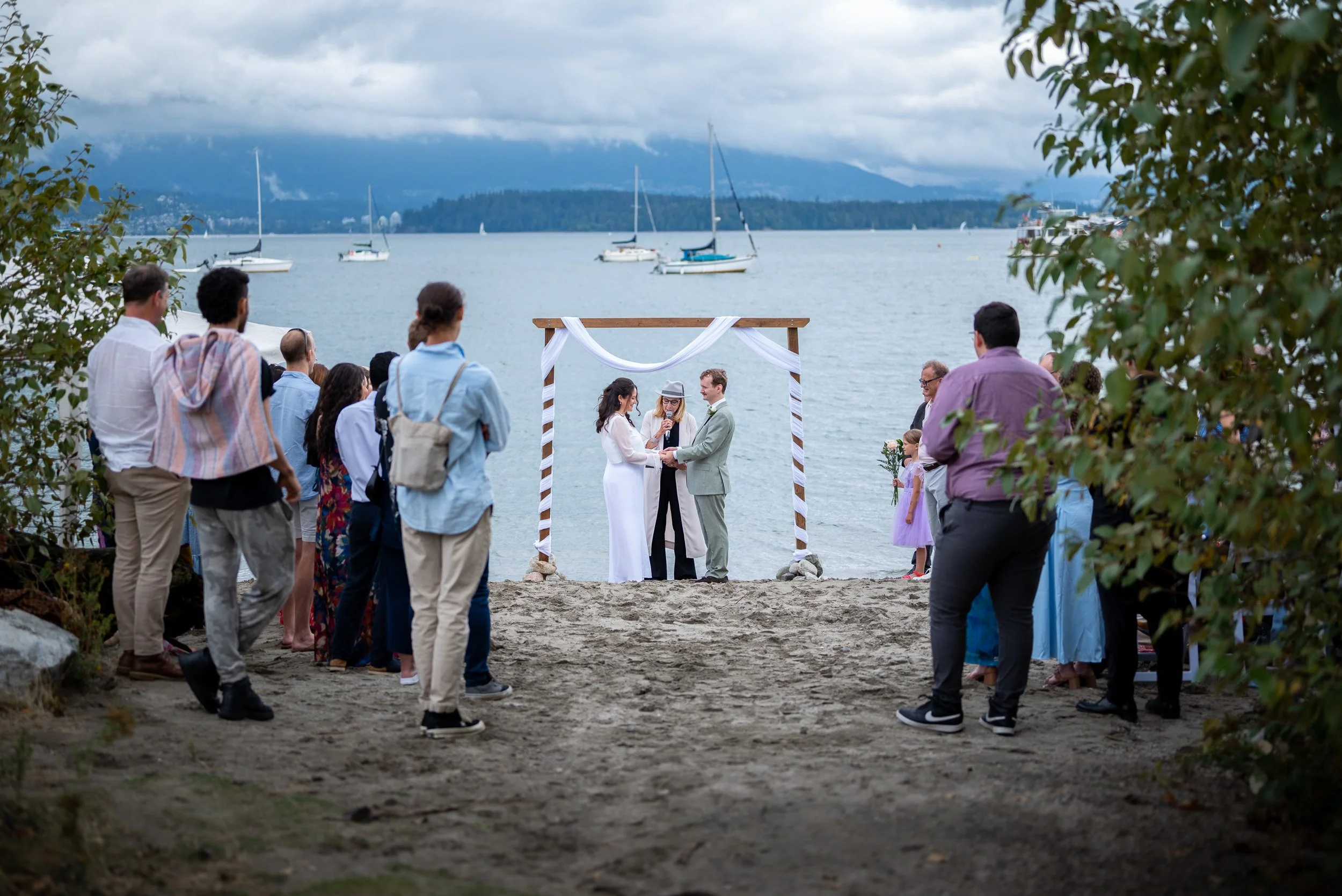 A group of people gathered on Jericho beach in Vancouver for a wedding ceremony, with an officiant and bride and groom under a wooden arch, overlooking a lake with sailboats and mountains in the background.