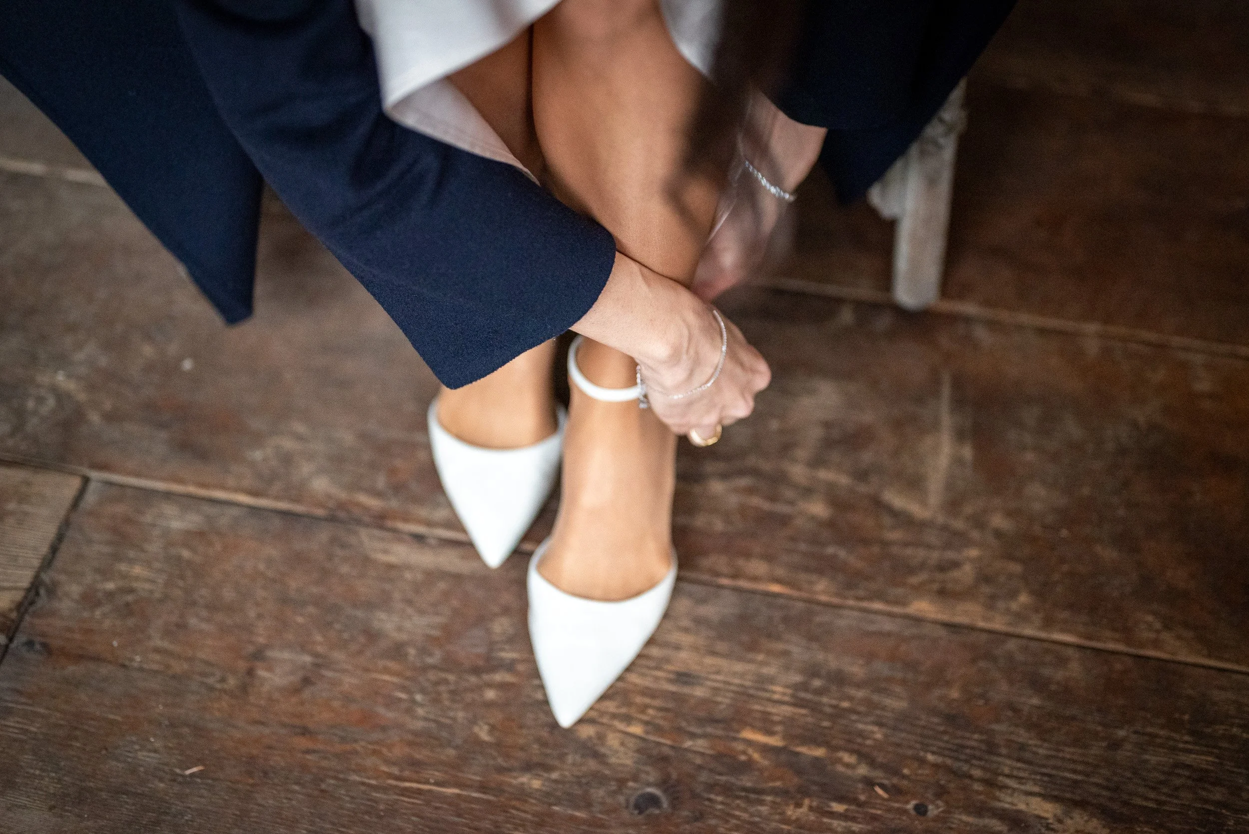 A getting ready photo of a bride putting on white high heels is sitting on a wooden chair. They are holding their ankle with their hand, and their foot is resting on the floor. The person has jewelry on their wrist, ankle, and finger.