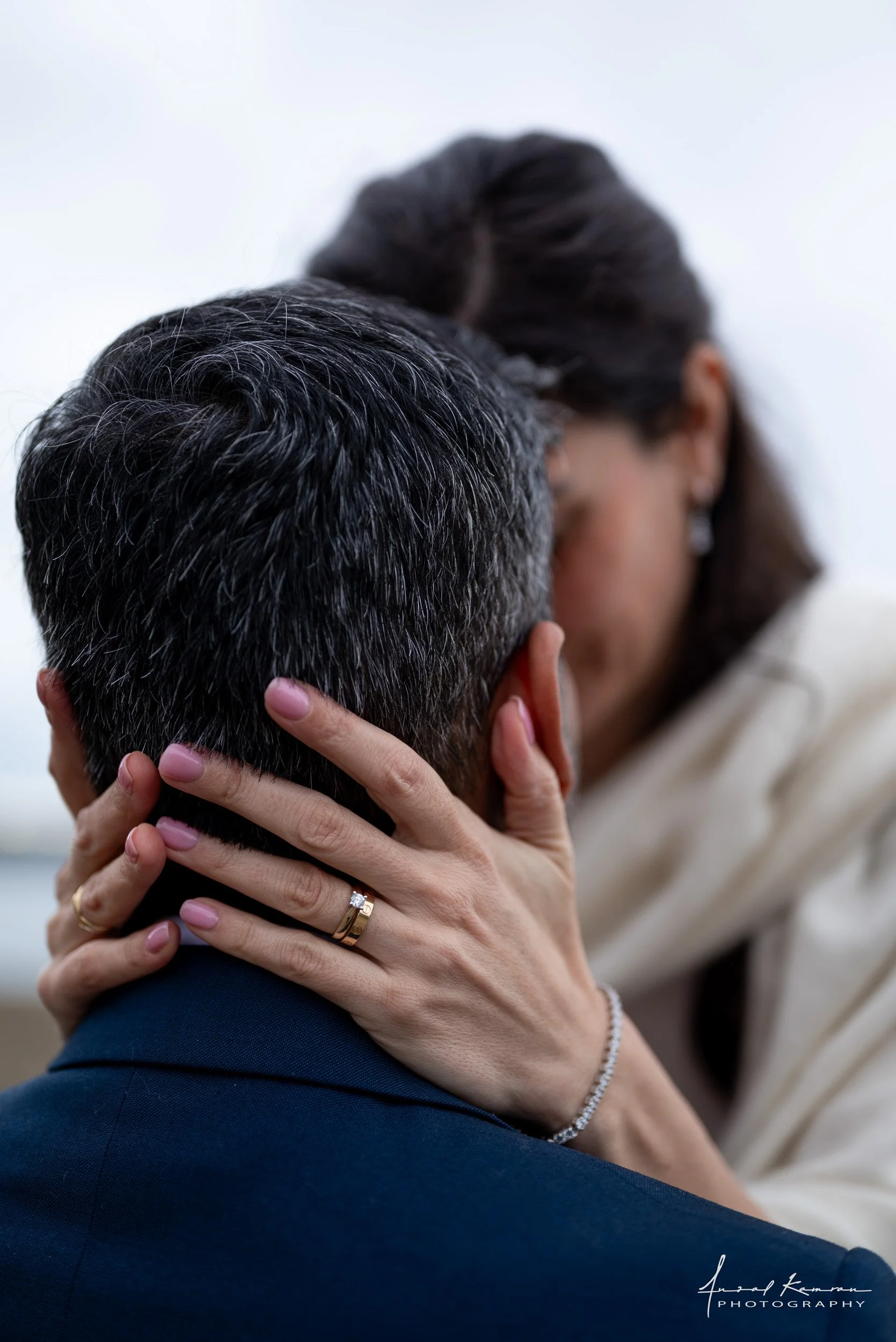 A woman and a man embracing in a close, intimate moment. The woman's hands are gently holding the man's face, and she is wearing rings and a bracelet. The man has short, dark hair with some gray strands, and the woman has shoulder-length dark hair. T