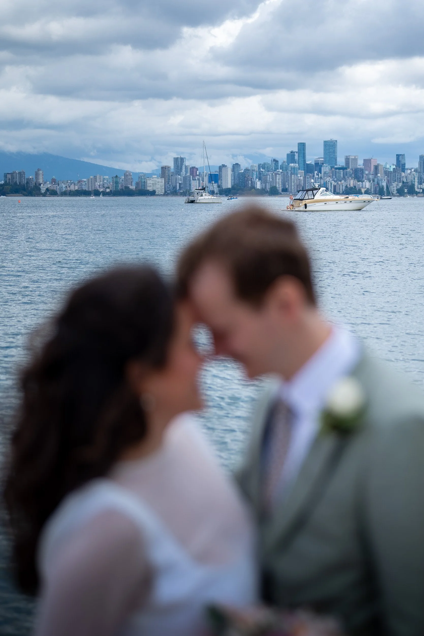 Blurred couple in wedding attire kissing with a city skyline and boats on the water in the background.