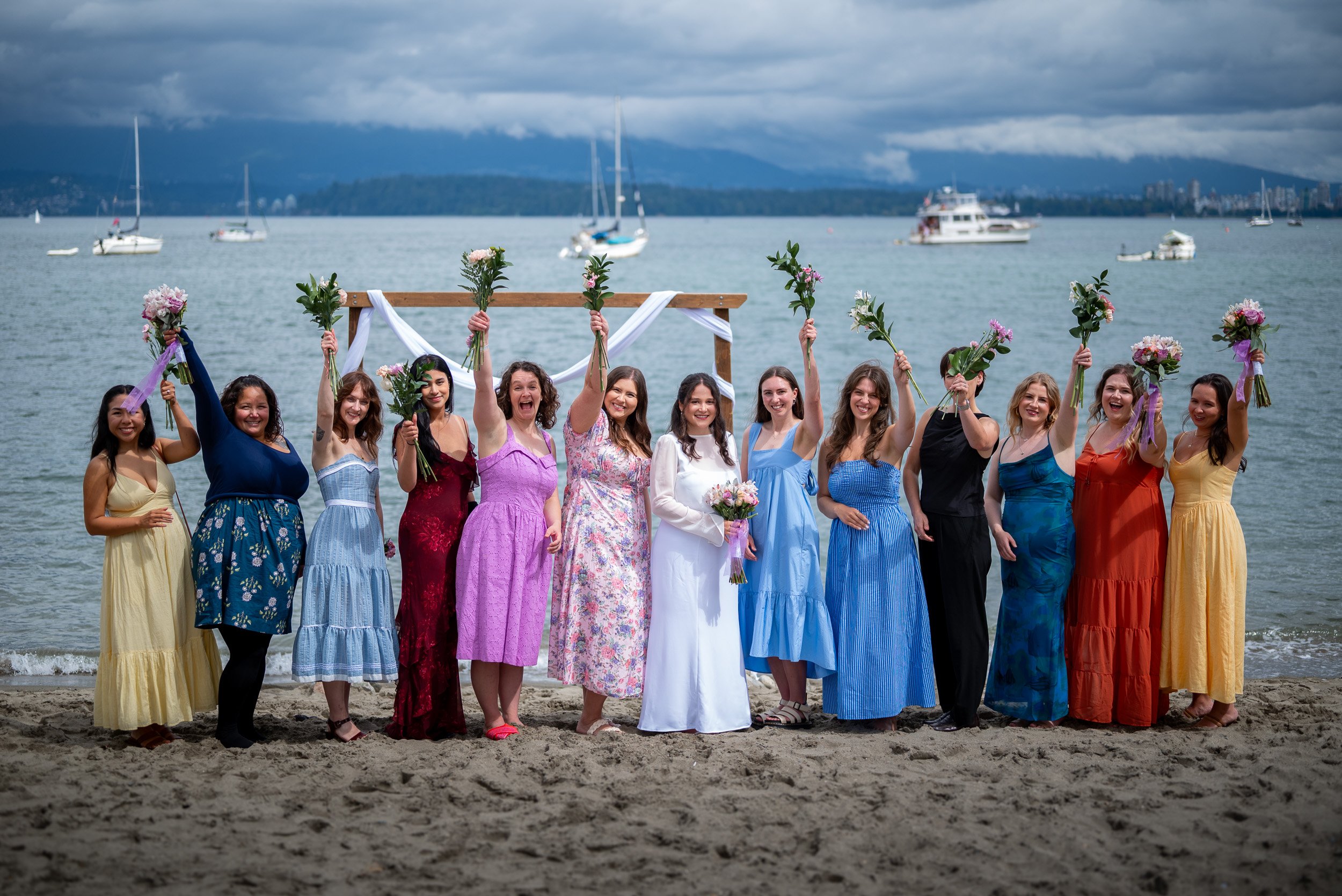 A group of women in colorful dresses holding bouquets of flowers at Jericho Beach wedding, with boats and a cloudy sky in the background.