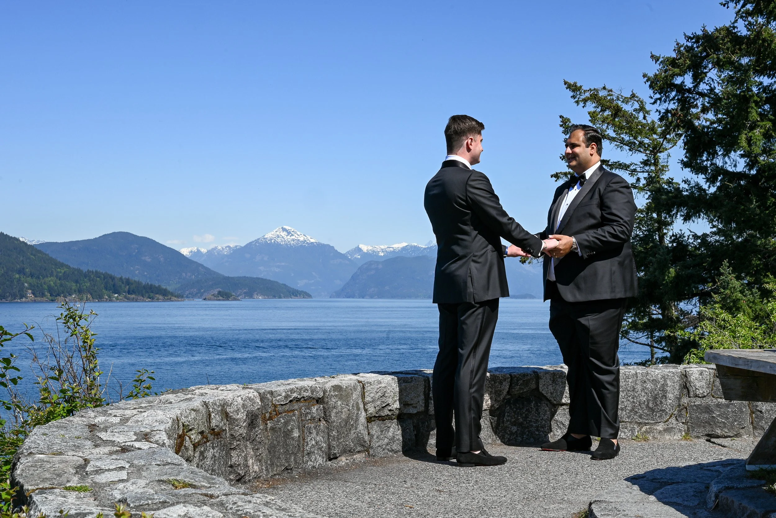 Two men in tuxedos getting married, holding hands and facing each other by a waterfront with mountains and snow-capped peaks in the background, under clear blue sky at Whytecliff Park in West Vancouver.
