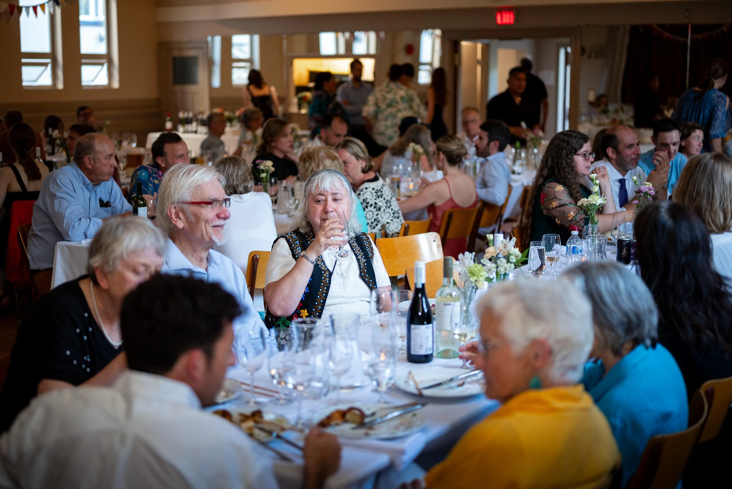 People seated at long dining tables decorated with flower arrangements, enjoying a gathering or celebration in a warmly lit room with multiple windows.