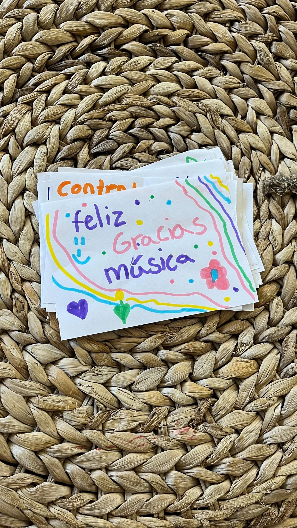 Colorful handwritten note that says 'feliz Gracias música' with decorative hearts, smiley face, flower, and colorful lines, placed on a woven straw surface.