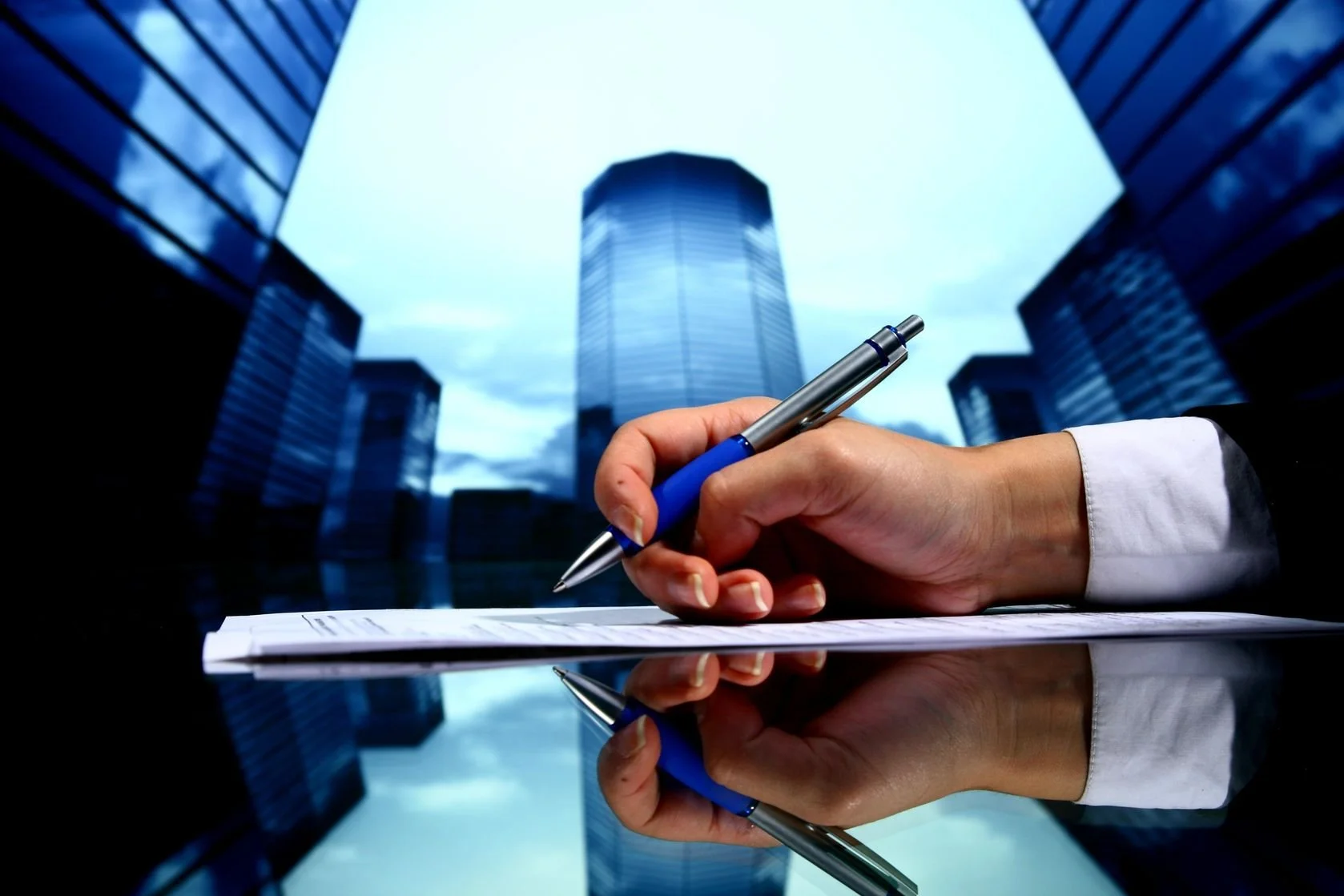 Persona firmando un documento sobre una mesa reflejante con edificios modernos de fondo.
