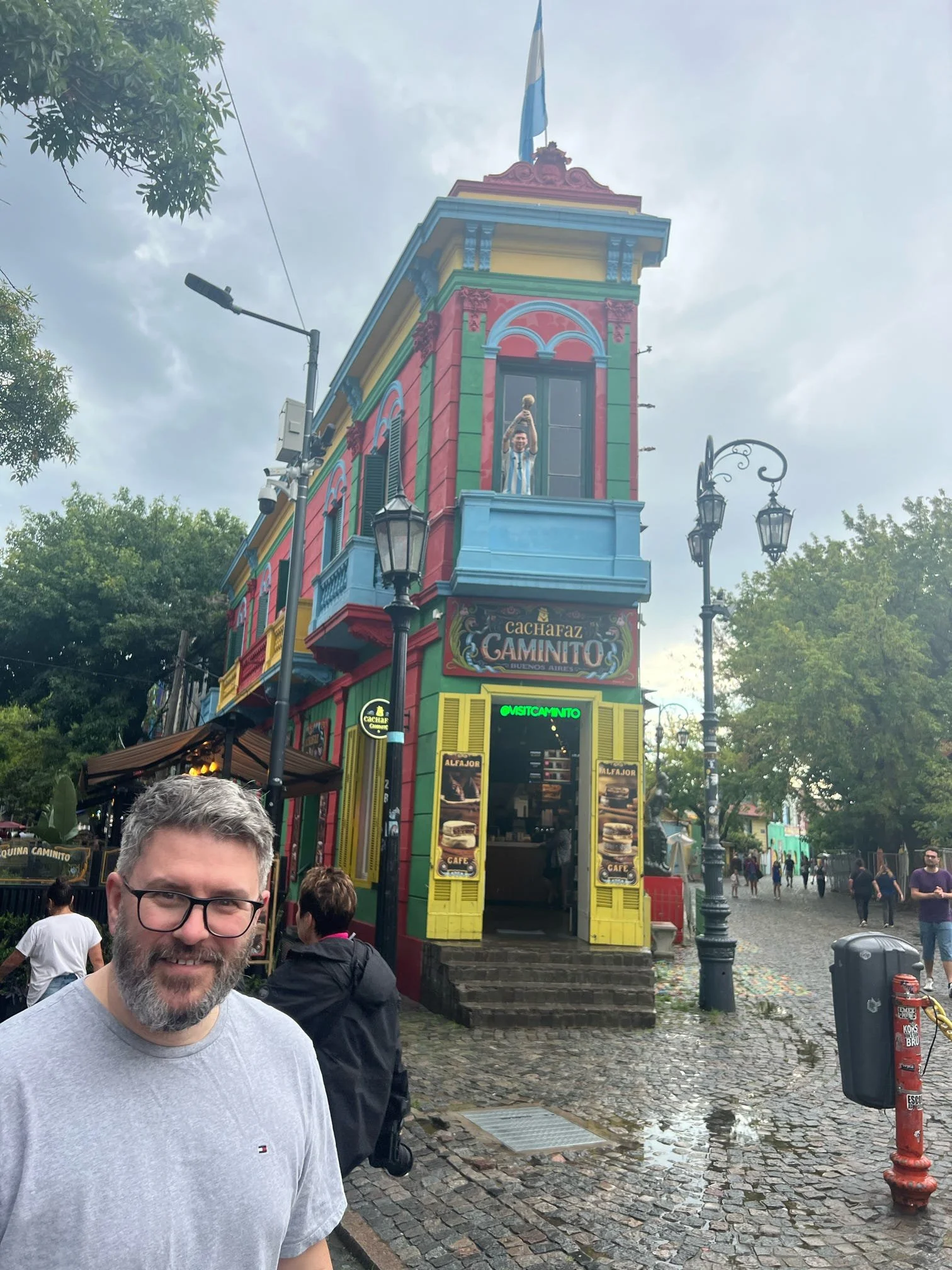 A man with glasses, a beard, and a gray t-shirt standing in front of a colorful building with pink, green, yellow, and blue accents in Buenos Aires, Argentina. The building has a sign that reads 'Cachafaz Caminito' and a person in the window holding 