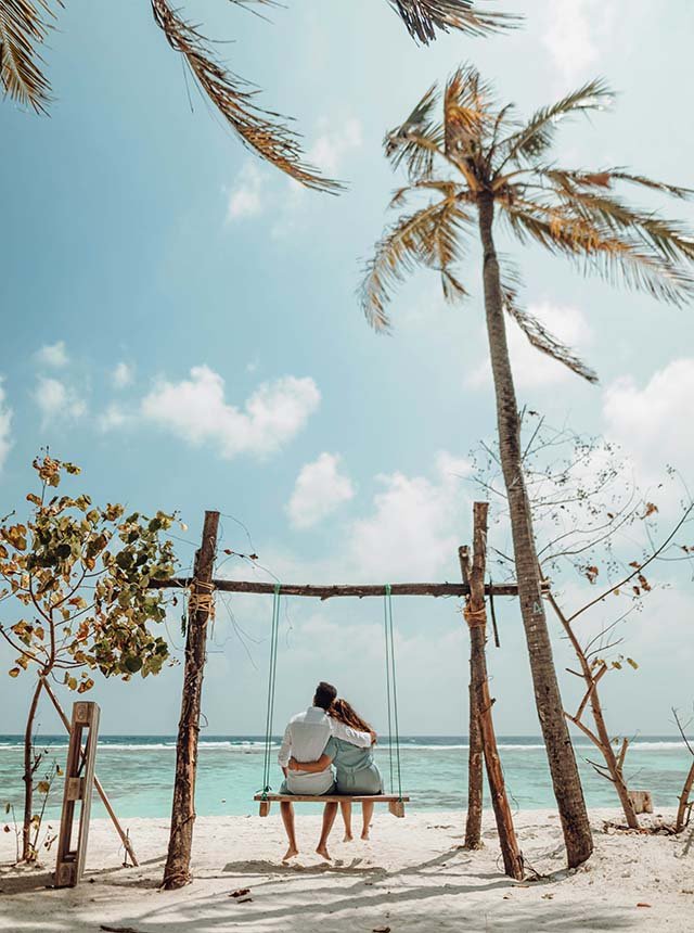 A couple sitting on a swing hanging from a wooden frame on a sandy beach, facing the ocean with palm trees overhead and a partly cloudy sky.