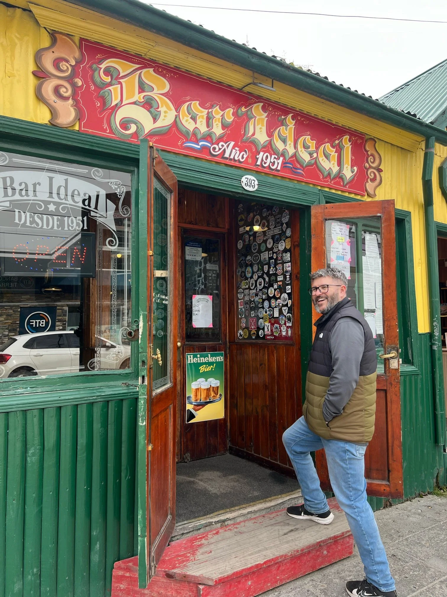 Man standing on a small red wooden porch outside a colorful restaurant with a red sign that says 'Bar'. The man is wearing glasses, a black vest over a gray hoodie, and jeans. The restaurant has a yellow and green exterior with a glass door and windo