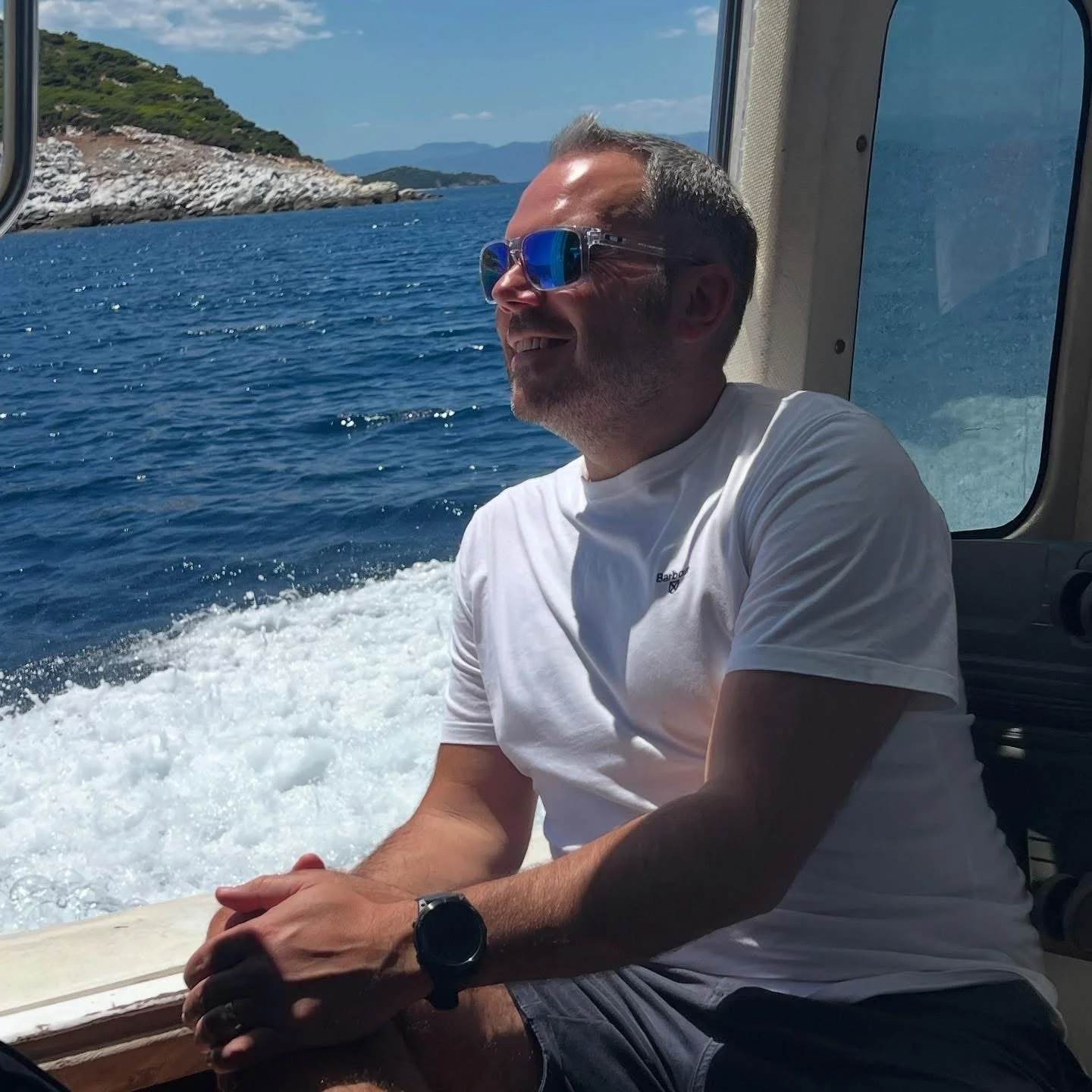 A man wearing sunglasses and a white t-shirt sitting on a boat, smiling while enjoying the water and a scenic coastline in the background.