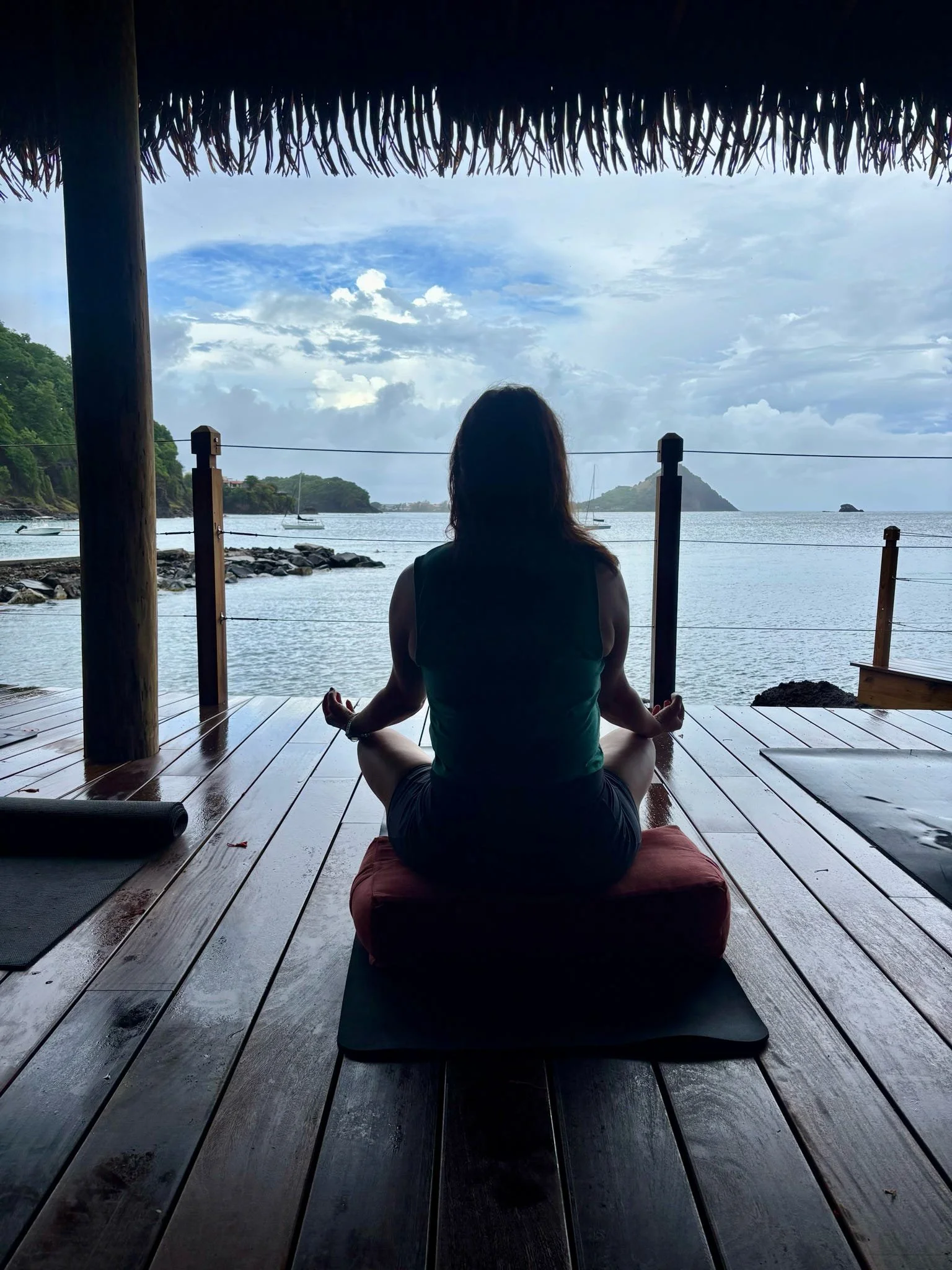A person practicing yoga in a seated meditation pose on a mat outdoors, overlooking a body of water with boats and islands in the distance, under a thatched roof