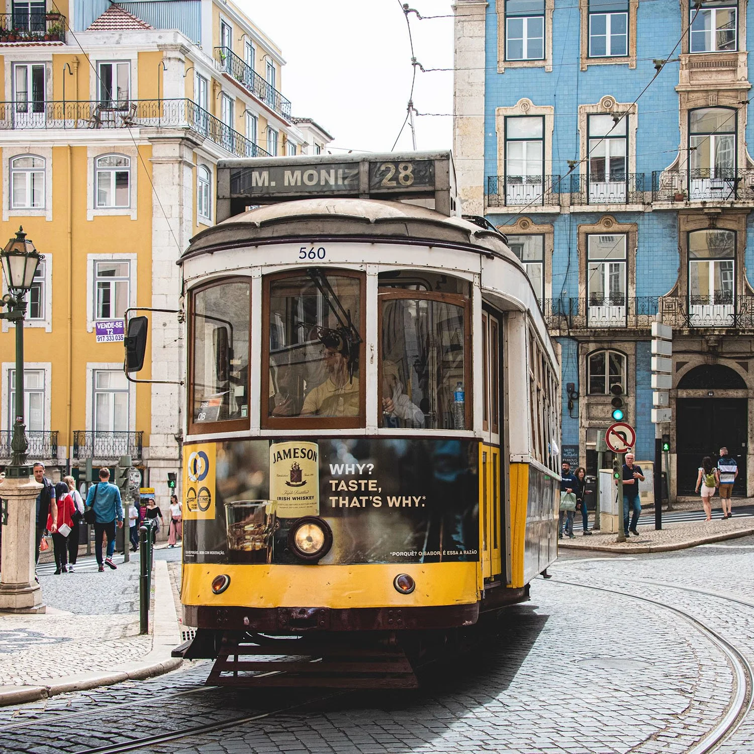 A yellow and white tram on cobblestone street with colorful European-style buildings in the background. People are walking and waiting at the tram stop.