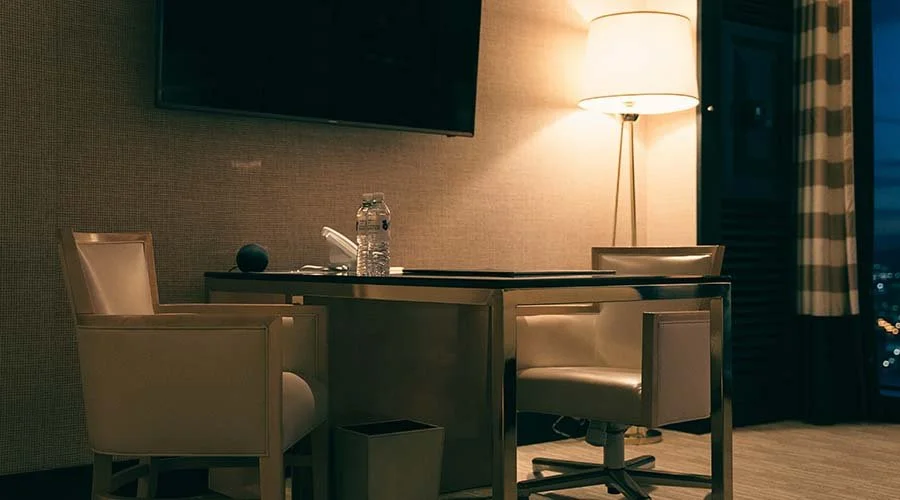 Hotel room with a table, two beige chairs, a water bottle, a black speaker, and a lamp near a window with curtains.