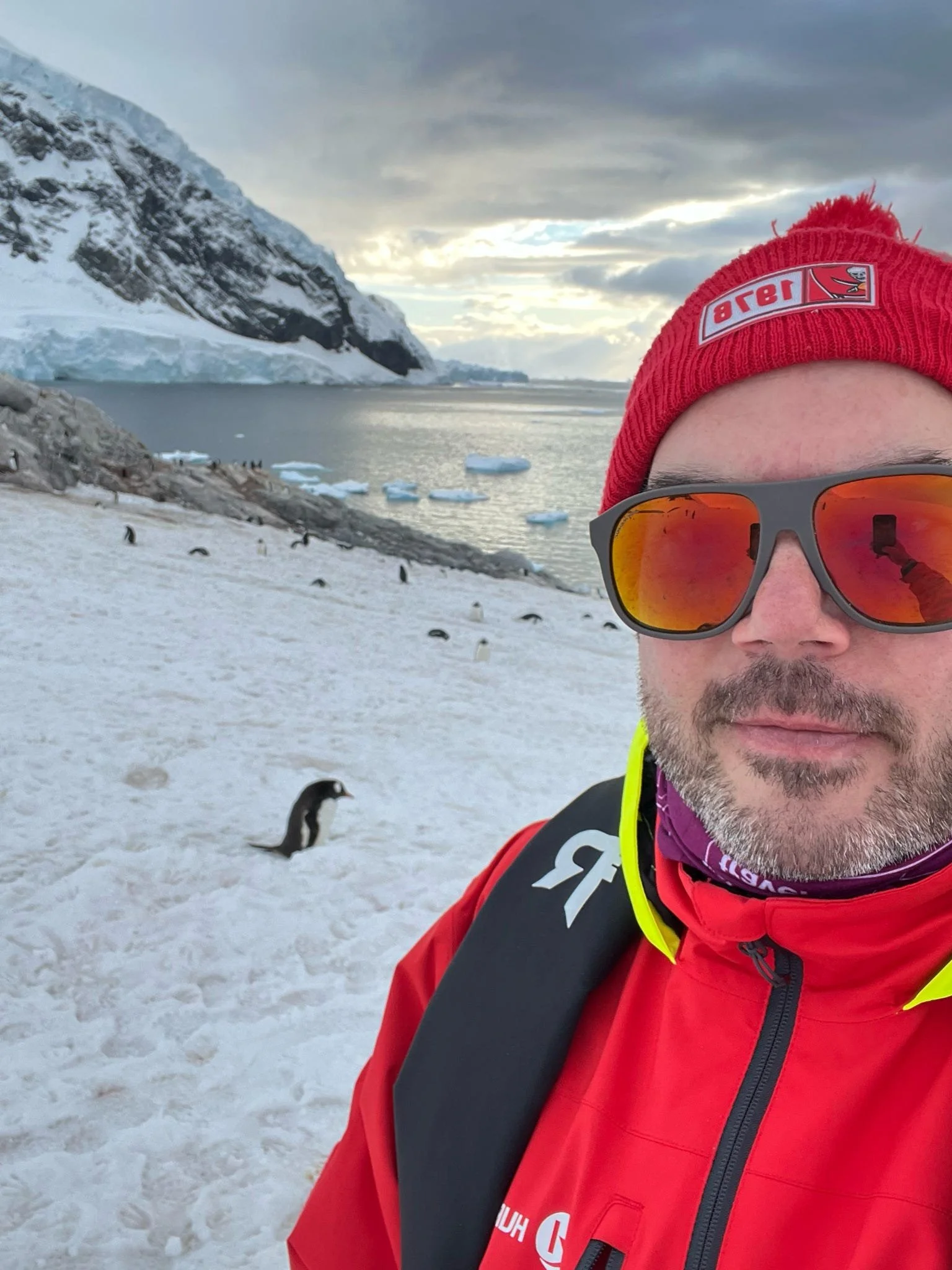 A person wearing red outdoor gear, sunglasses, and a red beanie with the Tampa Bay Lightning logo, standing on snow-covered terrain near a body of water with floating ice, mountains, and cloudy sky in the background.