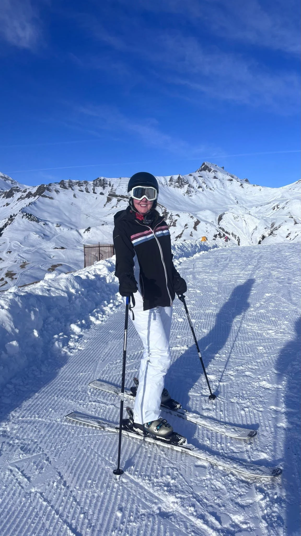 A woman dressed in winter ski gear, including a helmet, goggles, and a black jacket, standing on snowy ski slope with mountains and blue sky in the background.