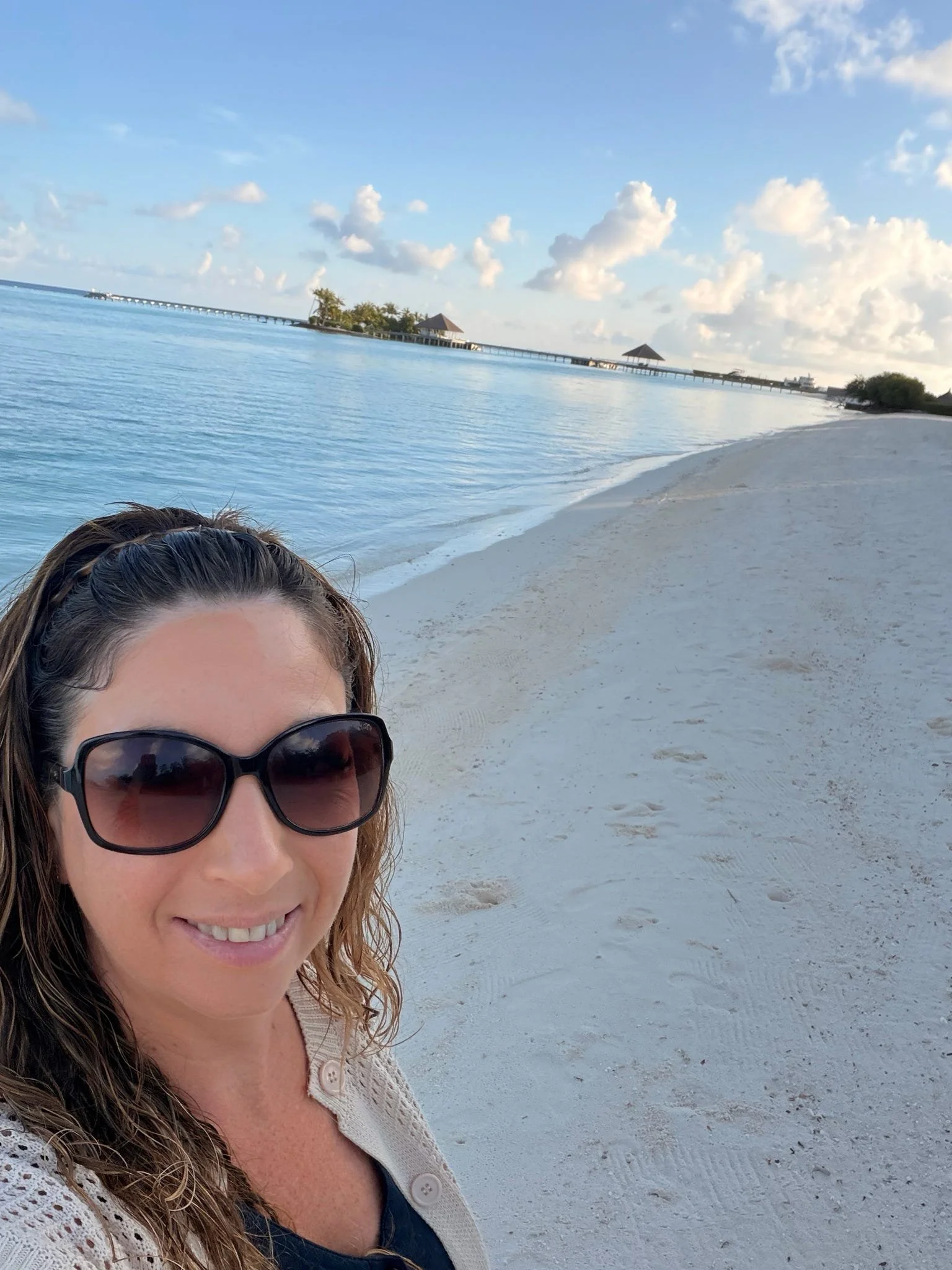 A smiling woman with long brown hair wearing sunglasses and a beige sweater, standing on a sandy beach with calm water and a pier with small huts and palm trees in the background.