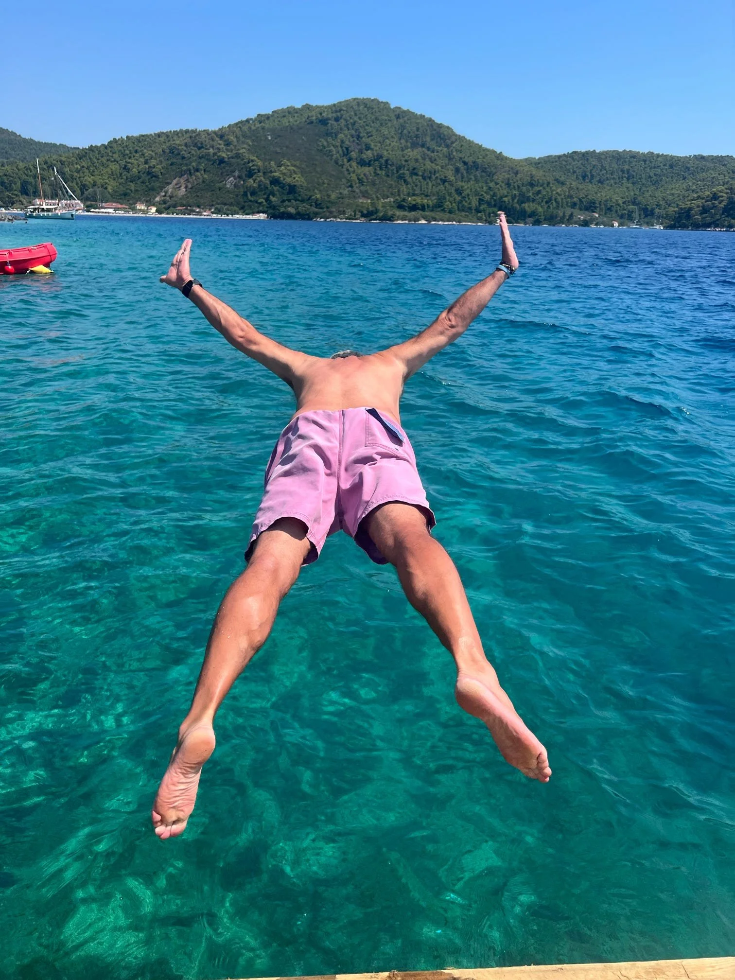A person in pink shorts is diving into clear blue water with a mountainous island in the background, and a sailboat is visible on the left side.