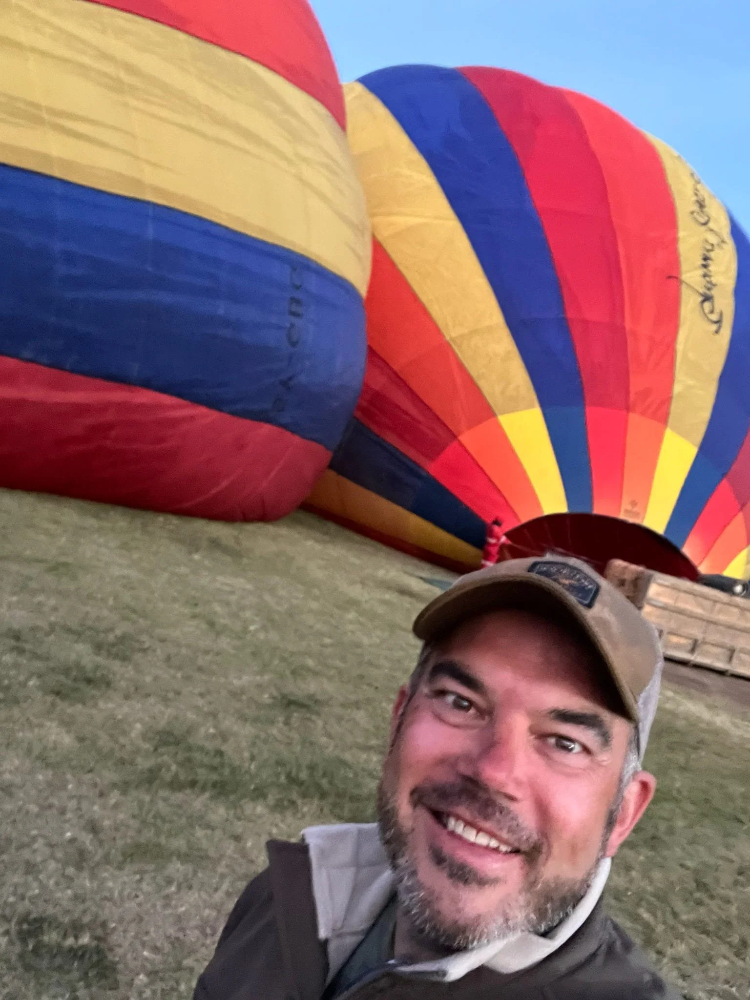 A man with a beard and glasses taking a selfie on the ground in front of deflated hot air balloons that are primarily yellow, red, and blue, with a clear sky in the background.