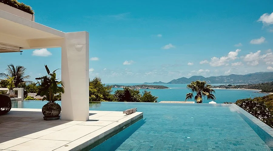 Luxury infinity pool overlooking a tropical bay with palm trees, hilly landscape, and blue sky with clouds.