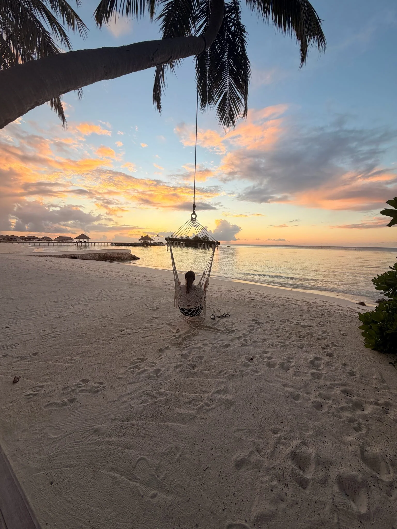 A person in a hammock on a sandy beach during sunset, with overwater bavilions in the distance, palm trees, and a cloudy sky with pink and orange hues.
