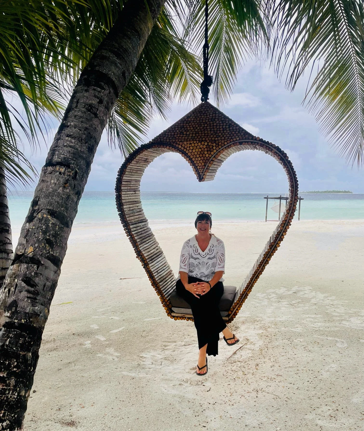 A woman sitting on a swing shaped like a heart made of wood logs, hanging from a palm tree on a sandy beach with ocean and cloudy sky in the background.