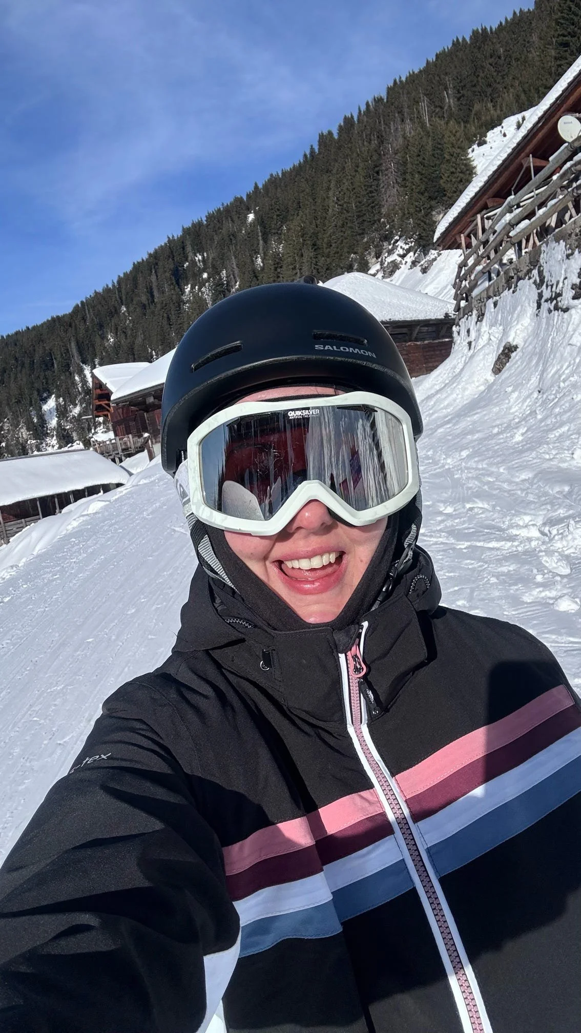 Person in ski gear with a helmet and goggles smiling for a selfie on snowy mountain slope with snow-covered houses and evergreen trees in the background.