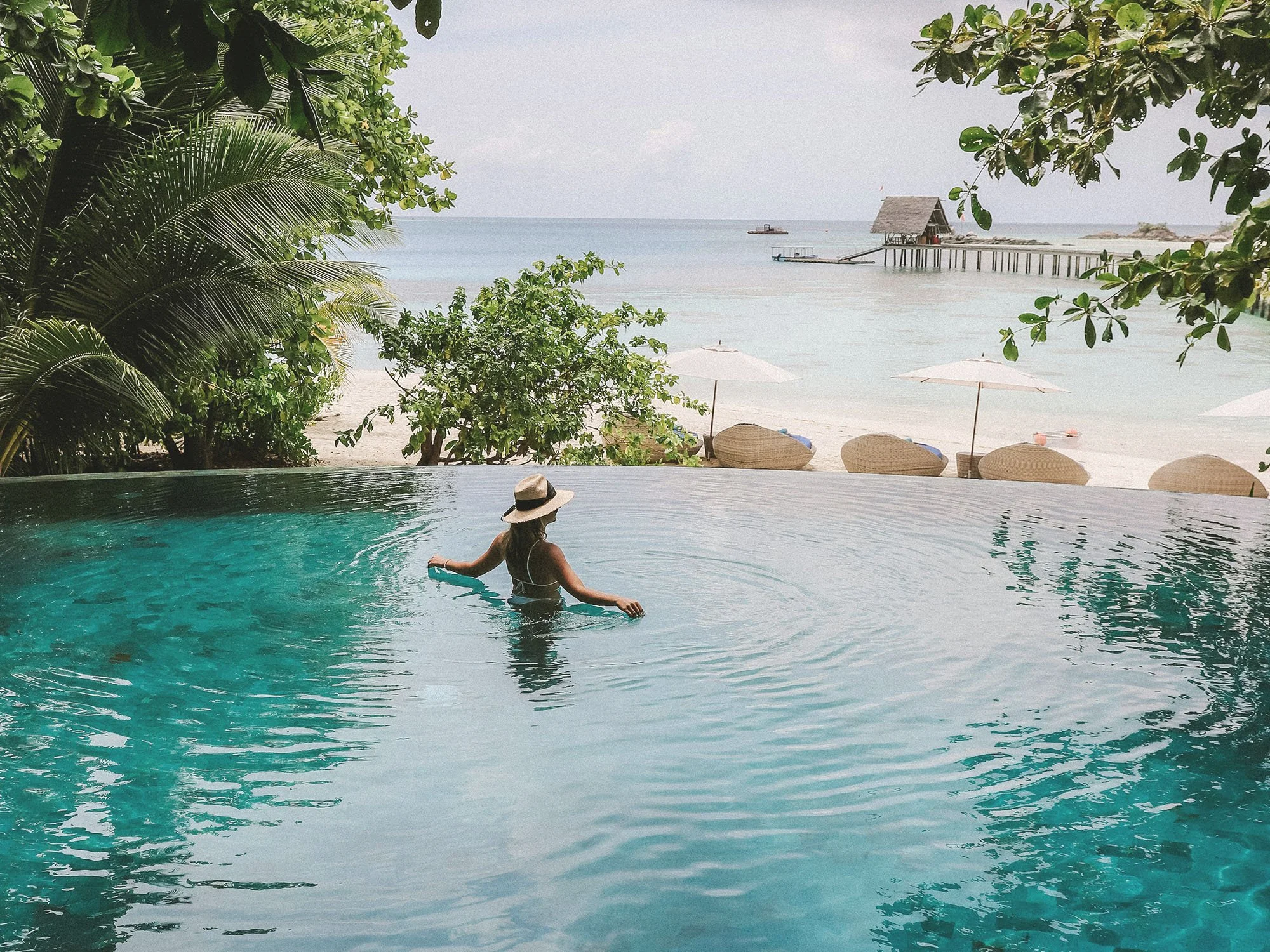 A woman sitting in a swimming pool near a beach, wearing a sun hat. In the background, a beach with umbrellas, lounge chairs, and a dock extends into the water, with trees framing the scene and a cloudy sky overhead.