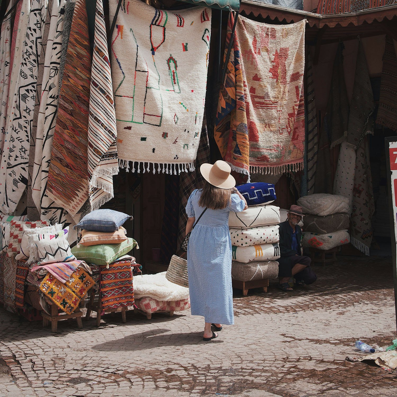A woman in a blue checkered dress and wide-brimmed hat looks at hanging and stacked traditional woven rugs and textiles outside a market stall. A man sits on some stacked cushions nearby. The scene is on a cobblestone street.