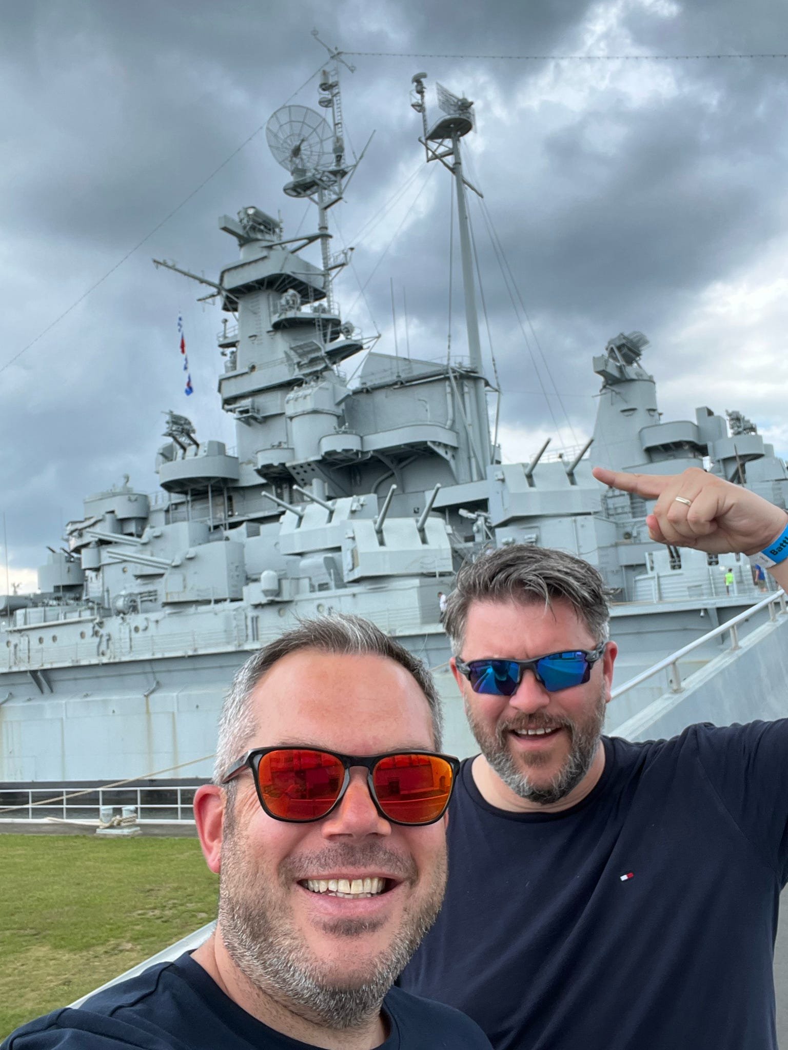 Two men taking a selfie in front of a large military ship with radar and communication antennas, with a cloudy sky overhead.