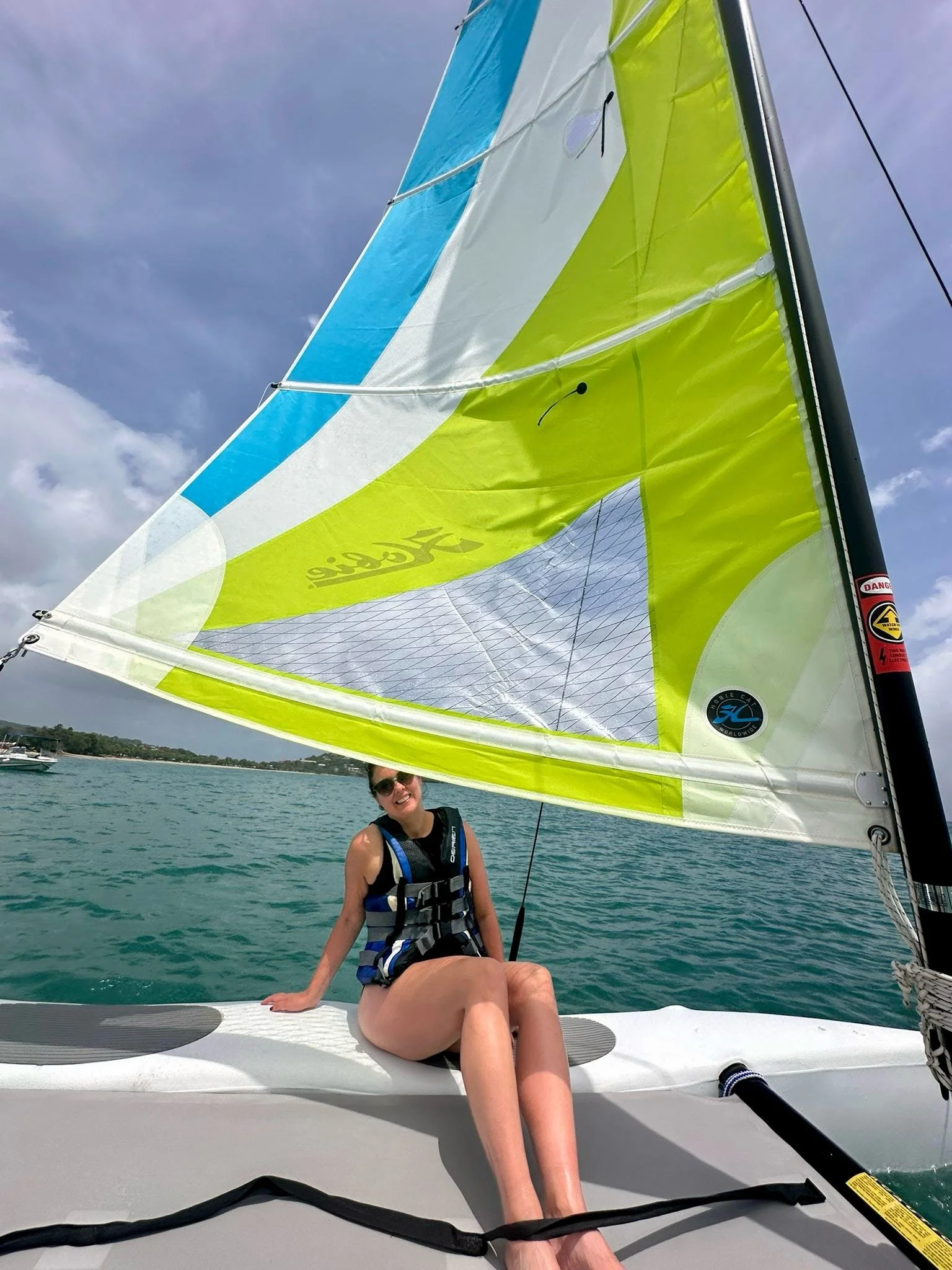 A woman sitting on the deck of a sailboat, smiling, with a large yellow and blue sail overhead, on a body of water under a partly cloudy sky.