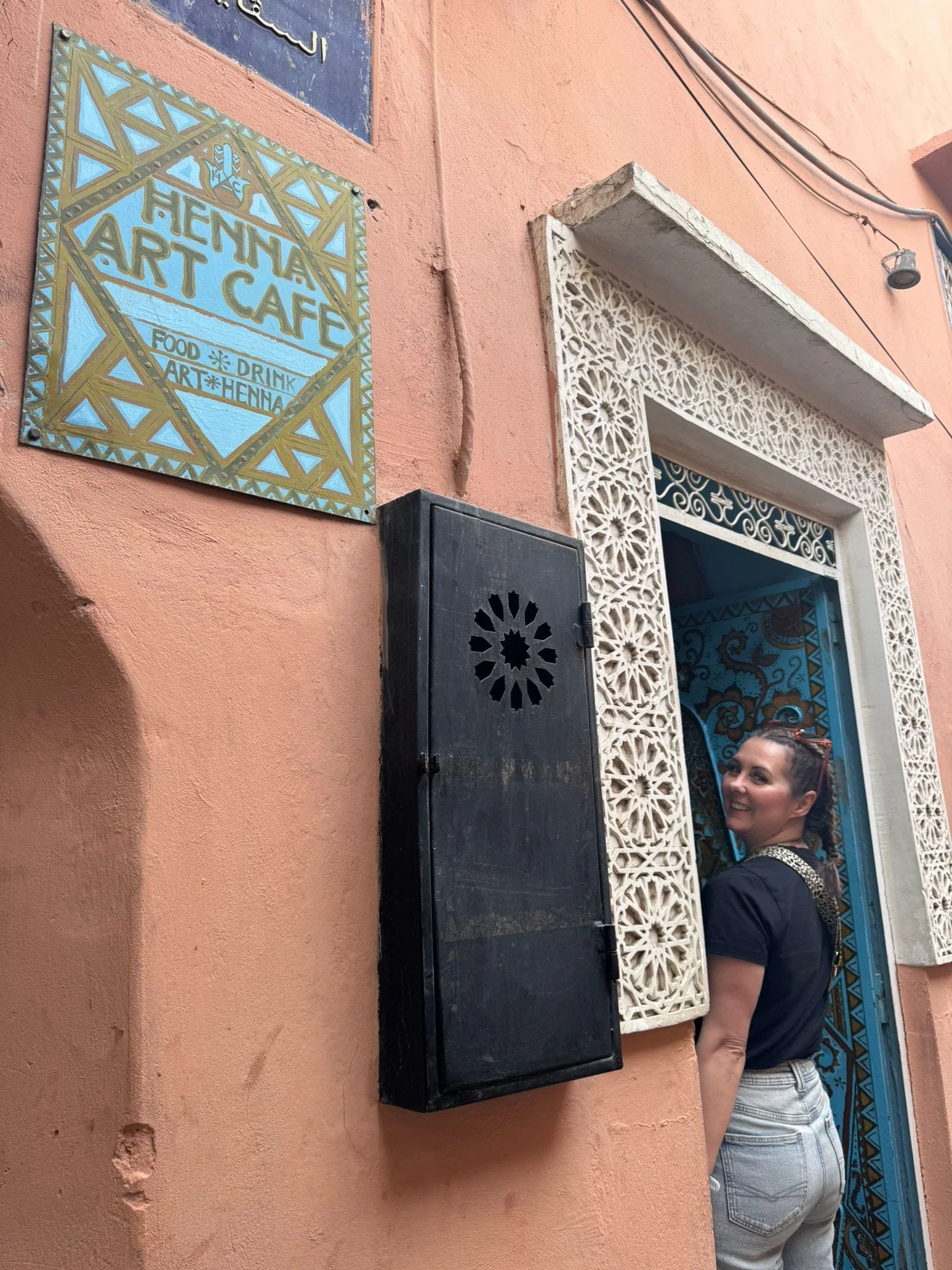 A woman with dark hair tied back, wearing a black t-shirt and light blue jeans, stands on a street near a building with light peach-colored walls, decorative white lattice window frame, and a sign for 'HENNA ART CAFE' that mentions food, drink, art, 