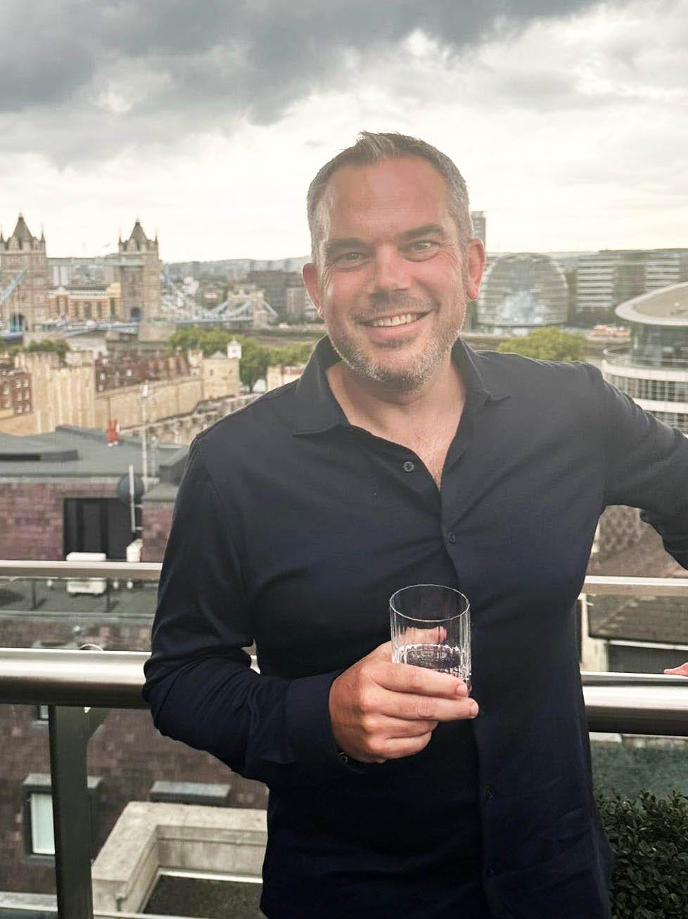 Smiling man with short hair and beard, wearing a black shirt, holding a glass, on a balcony with cityscape and landmarks in the background.
