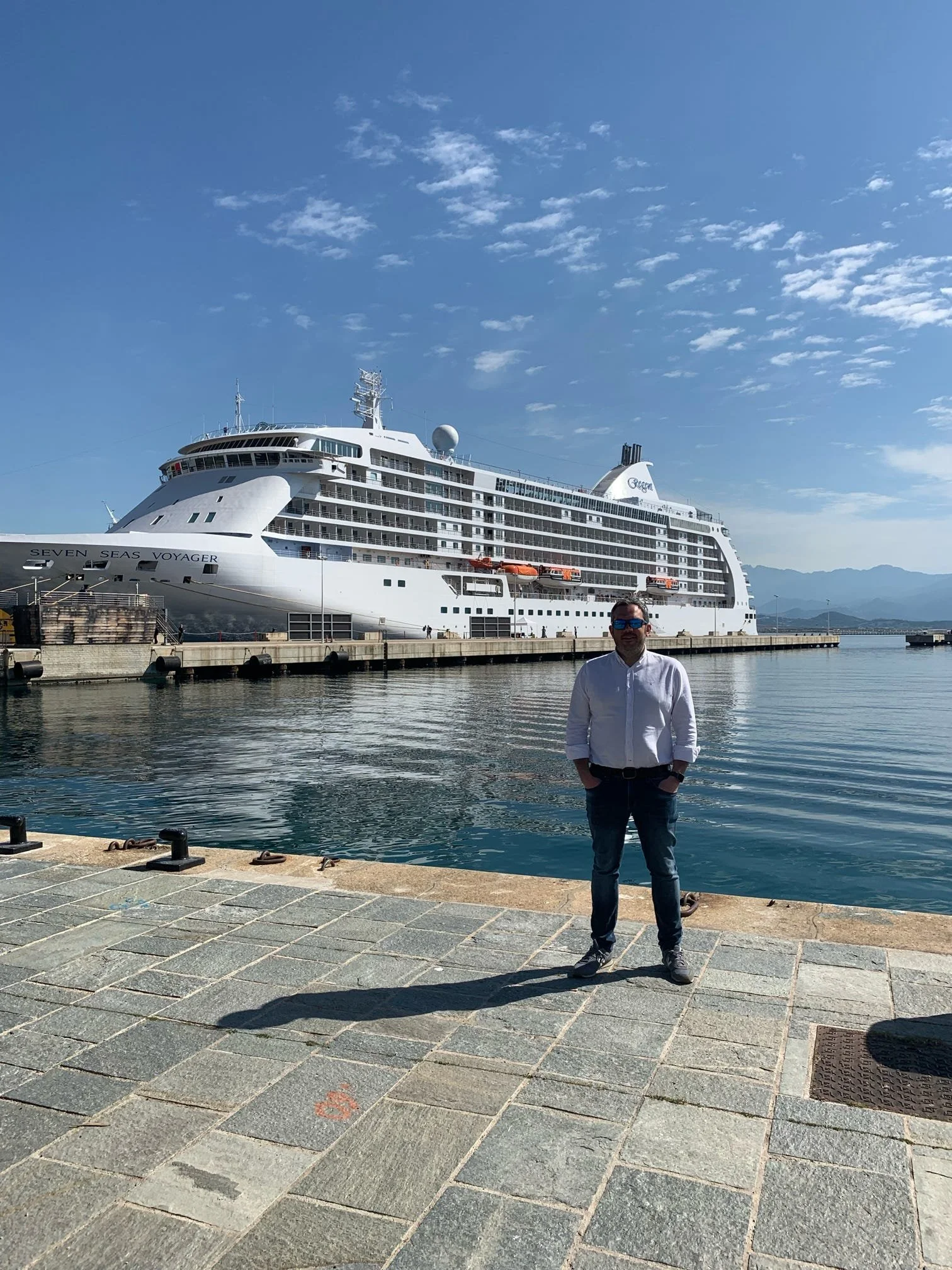 A man standing on a waterfront promenade with a large white cruise ship docked behind him. The sky is blue with scattered clouds, and there are mountains in the distance.