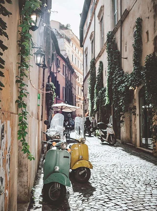 A narrow cobblestone street in a European city with pastel-colored buildings covered in green ivy. Several scooters are parked along the side, and a person is seen walking in the distance under an umbrella shading an outdoor cafe.