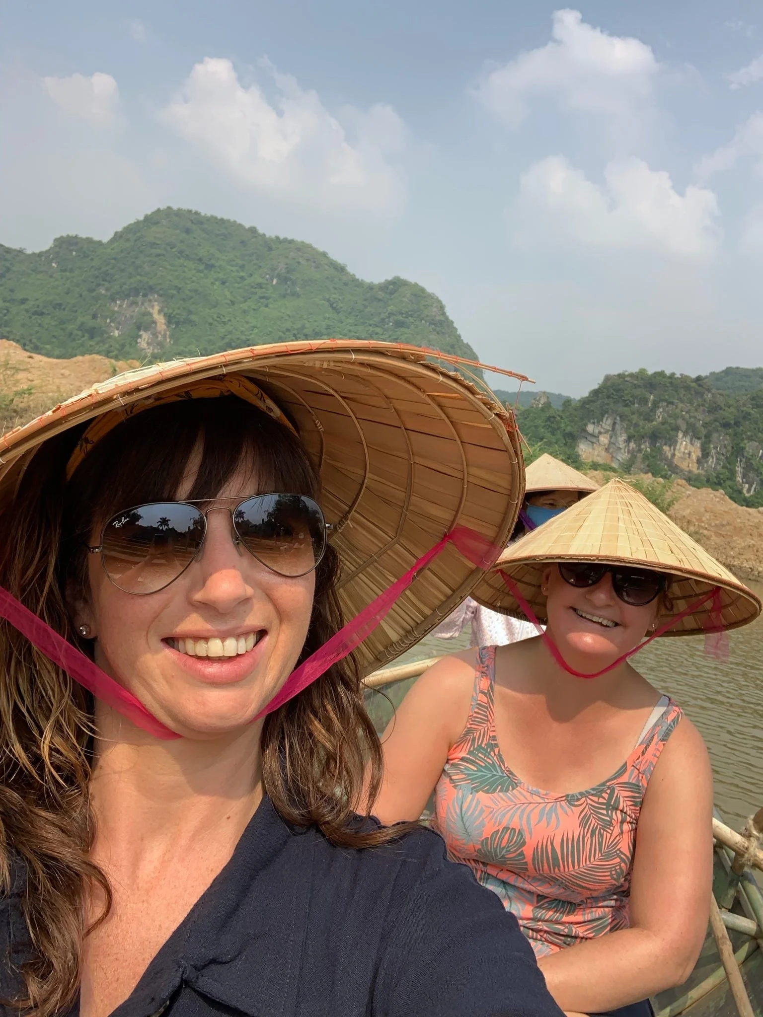 Two women wearing traditional Asian straw hats and sunglasses are taking a selfie in a scenic outdoor location with mountains, sky, and other people in the background.