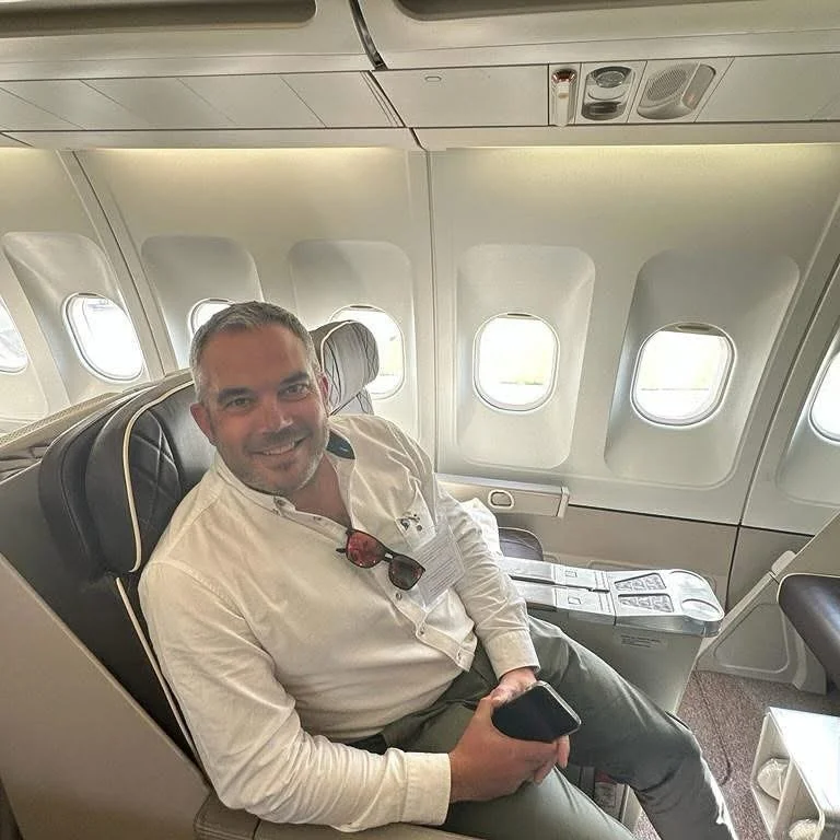 A man sitting in an airplane seat next to the window, smiling, holding a phone, with sunglasses hanging from his shirt collar.