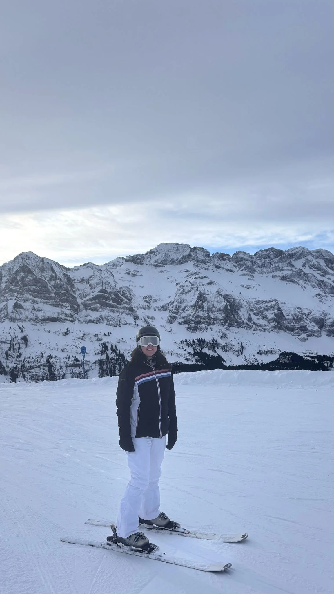 Person standing on skis in snowy mountain landscape, wearing a black helmet and goggles, black jacket with white and red accents, white pants, with snow-covered mountains in the background.