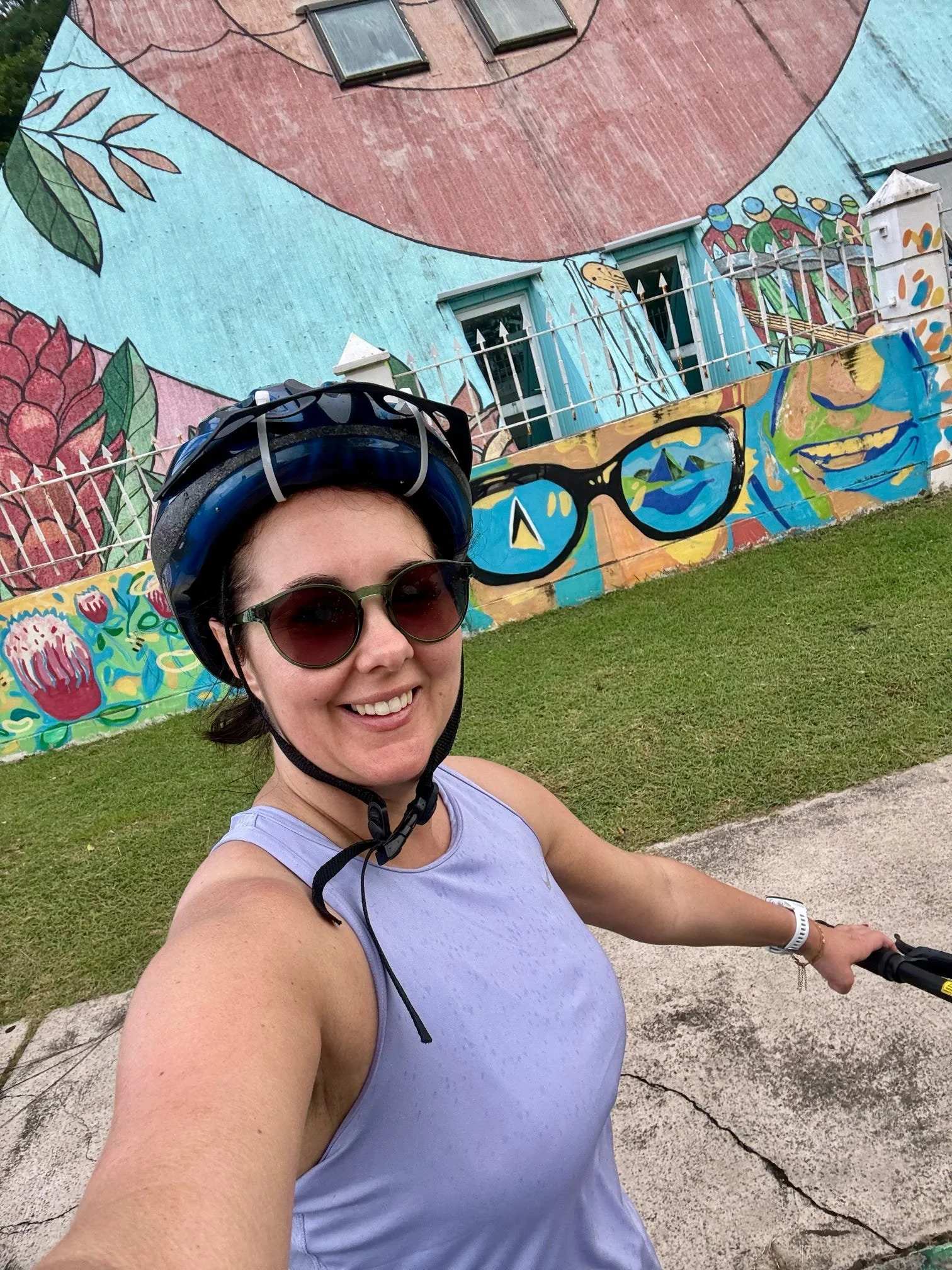 Close-up of a woman wearing sunglasses, a bicycle helmet, and a lavender tank top, smiling while riding a bike in front of a colorful mural on a building with trees in the background.