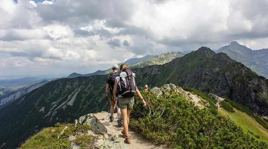 A group of hikers walking along a narrow mountain trail with lush green hills and rugged peaks under a cloudy sky.