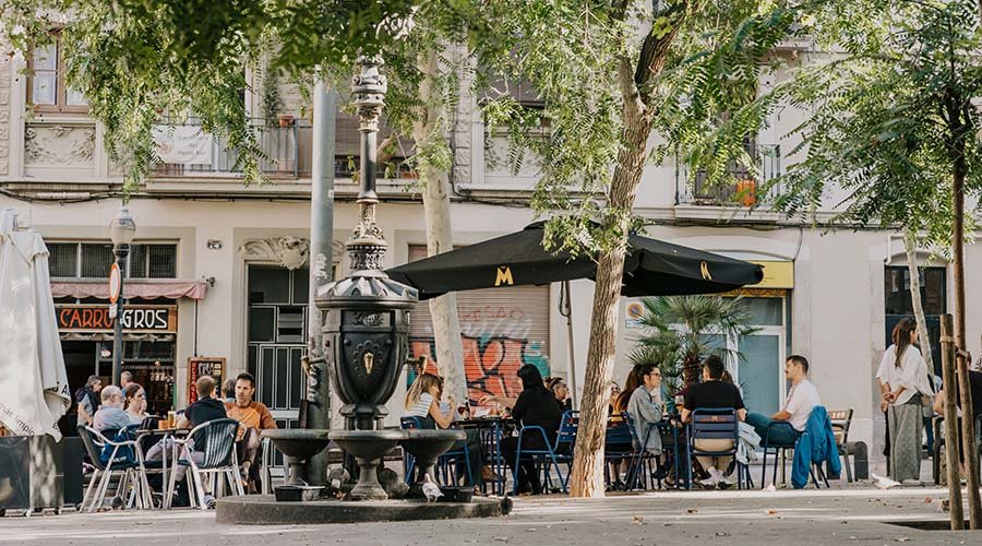 Outdoor café on a city street with people sitting at tables under umbrellas and a large tree in the foreground.