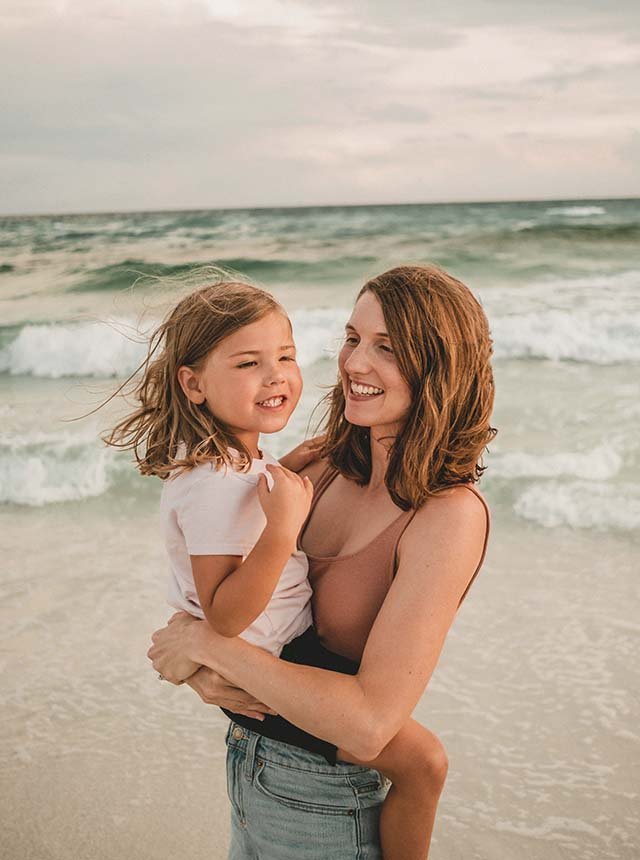 A woman and a young girl smiling and hugging on the beach, with the ocean waves in the background.