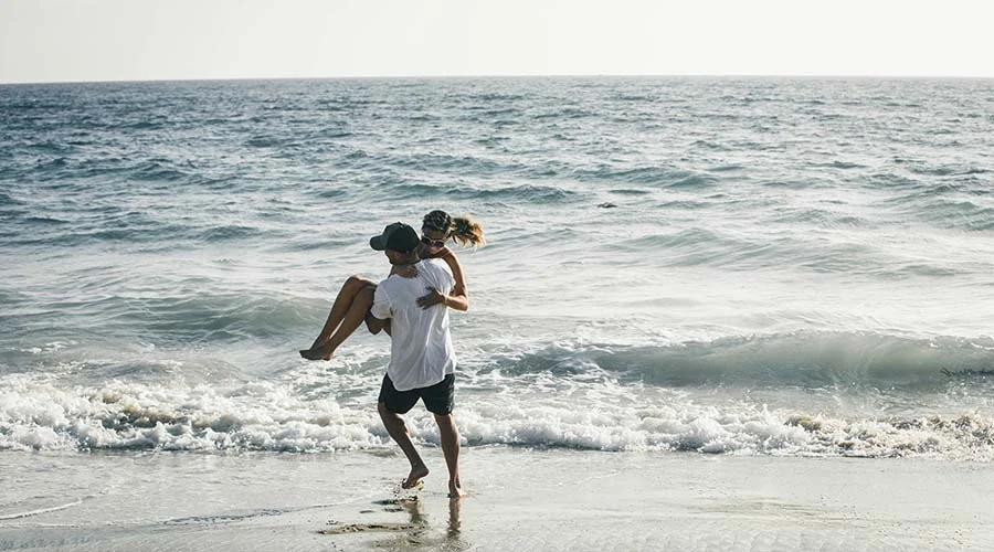 A person carrying a child on their back along the shoreline of a beach with gentle waves and an ocean in the background.