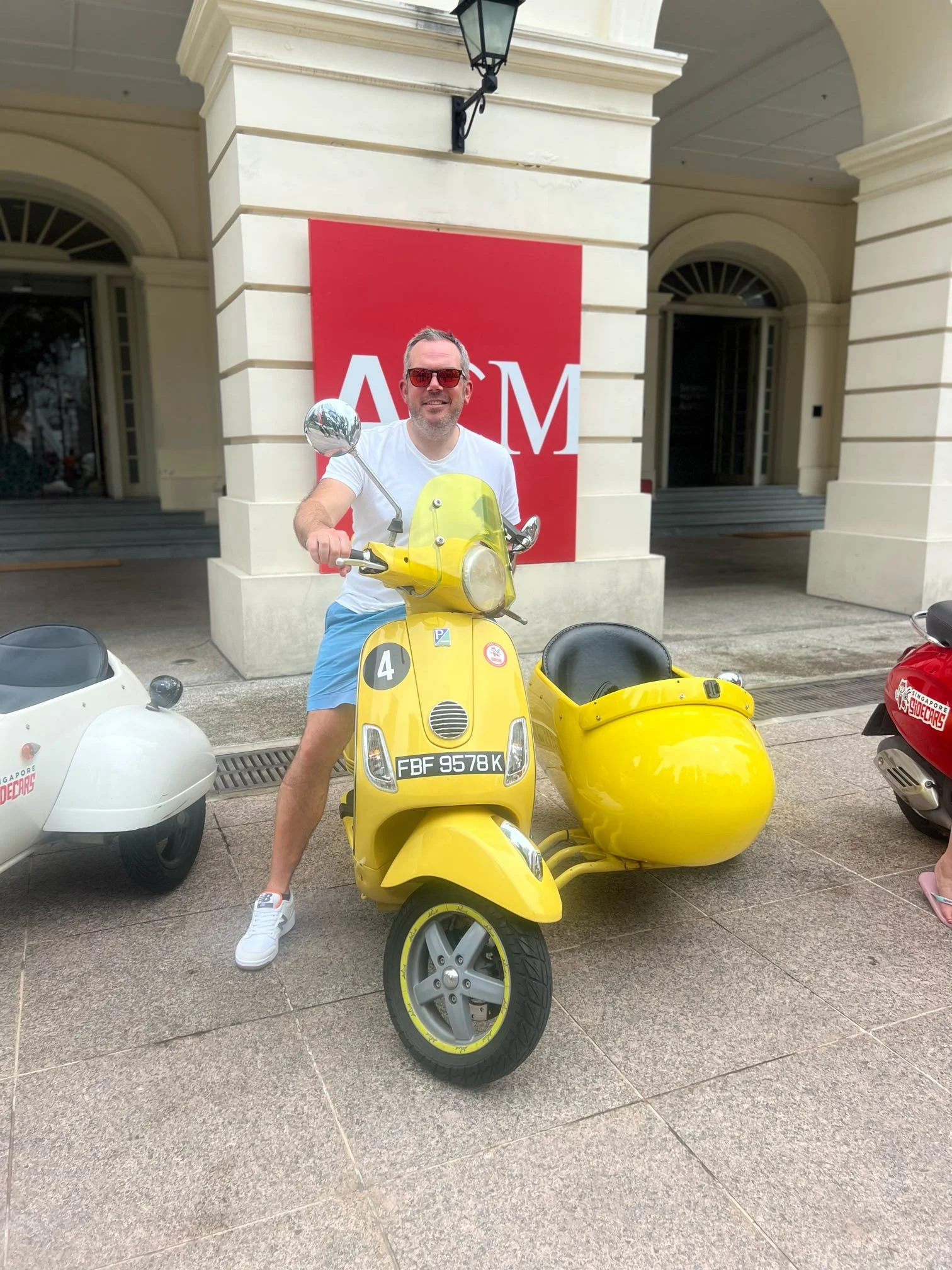 A man smiling and wearing sunglasses is standing next to a yellow scooter with a sidecar. The scooter has the license plate FBF 9578 K and is parked outside a building with a red sign that has white letters, partially visible, spelling 'ASM'. Other s