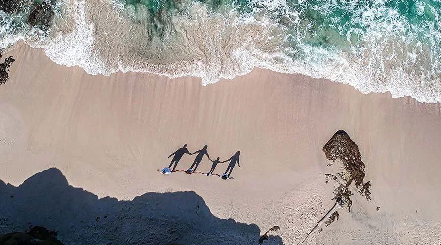 Aerial view of four people holding hands and walking on the beach with waves approaching from the ocean, and a large rock formation on the right side.