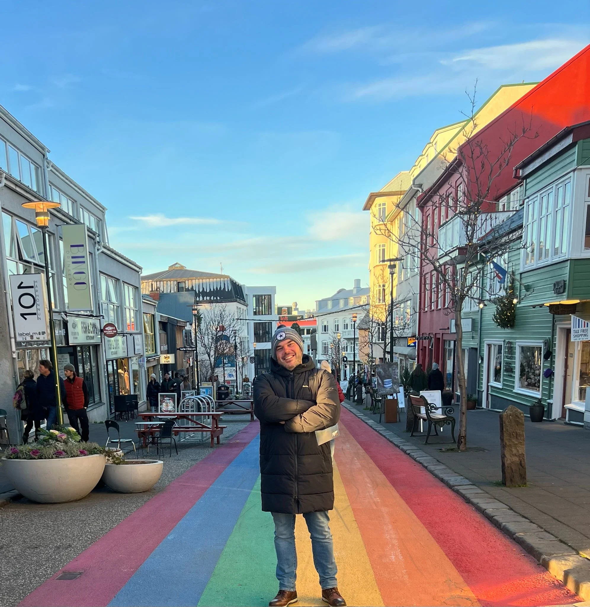 A man standing on a colorful rainbow-patterned crosswalk in an urban shopping district, smiling and wearing a winter coat and beanie, with shops and pedestrians in the background.