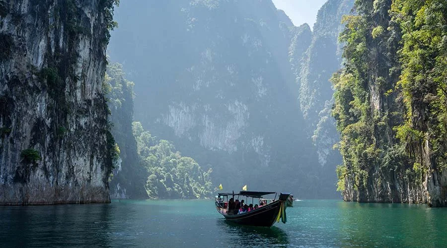 A boat with passengers floating through a narrow, scenic waterway flanked by tall, lush cliffs on either side.