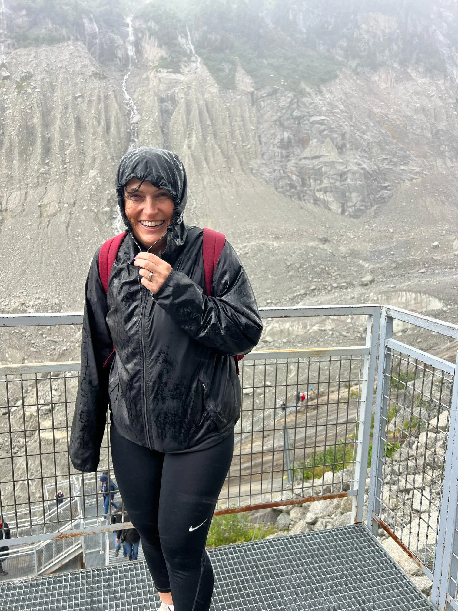 A woman smiling, wearing a black waterproof jacket with the hood up, standing on a metal staircase with rocky mountain terrain and waterfalls in the background.